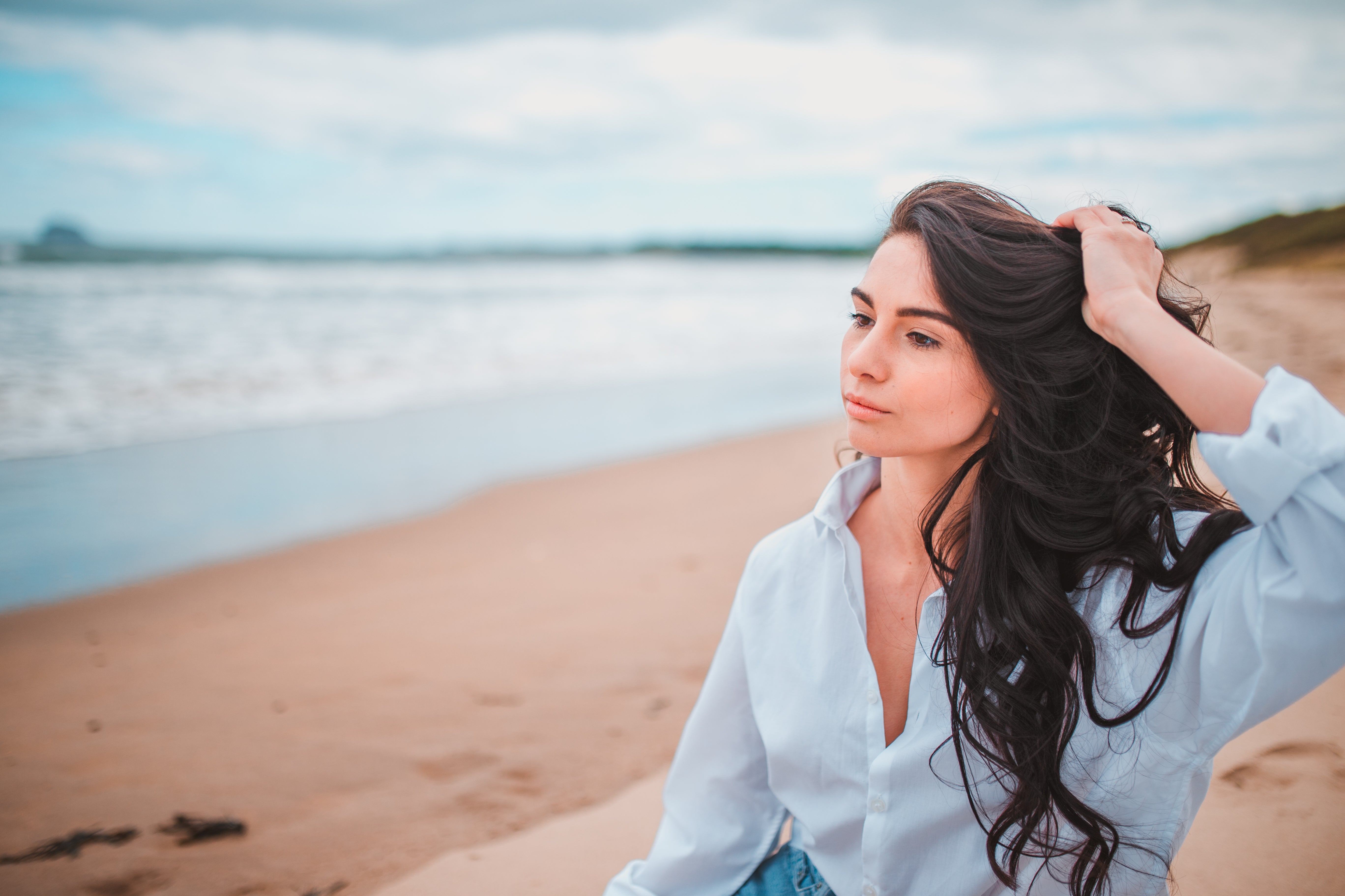 A female model is sitting on a beach looking out towards the sea. She has long, wavy, brown hair and has one hand placed on her head scrunching her waves. She is wearing a blue shirt and blue jeans. She has a neutral expression on her face.