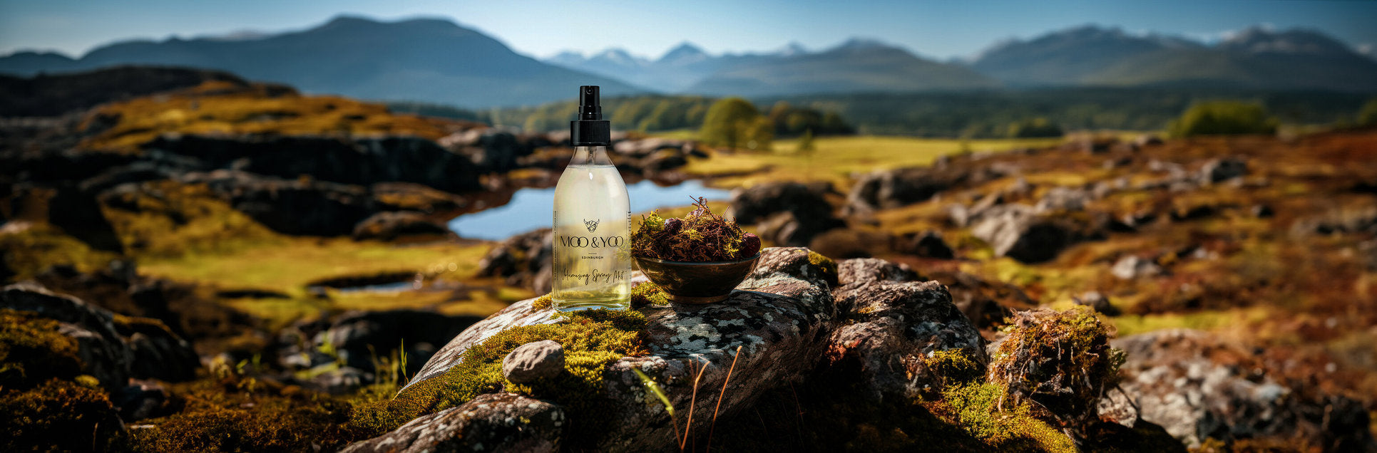 A bottle of Moo & Yoo Volumising Spray Mist sitting on a large rock which is covered in moss. The camera is focused on the bottle and there is a bowl full of moss sitting on the right of the bottle. There is a mountainous background with lots of greenery.