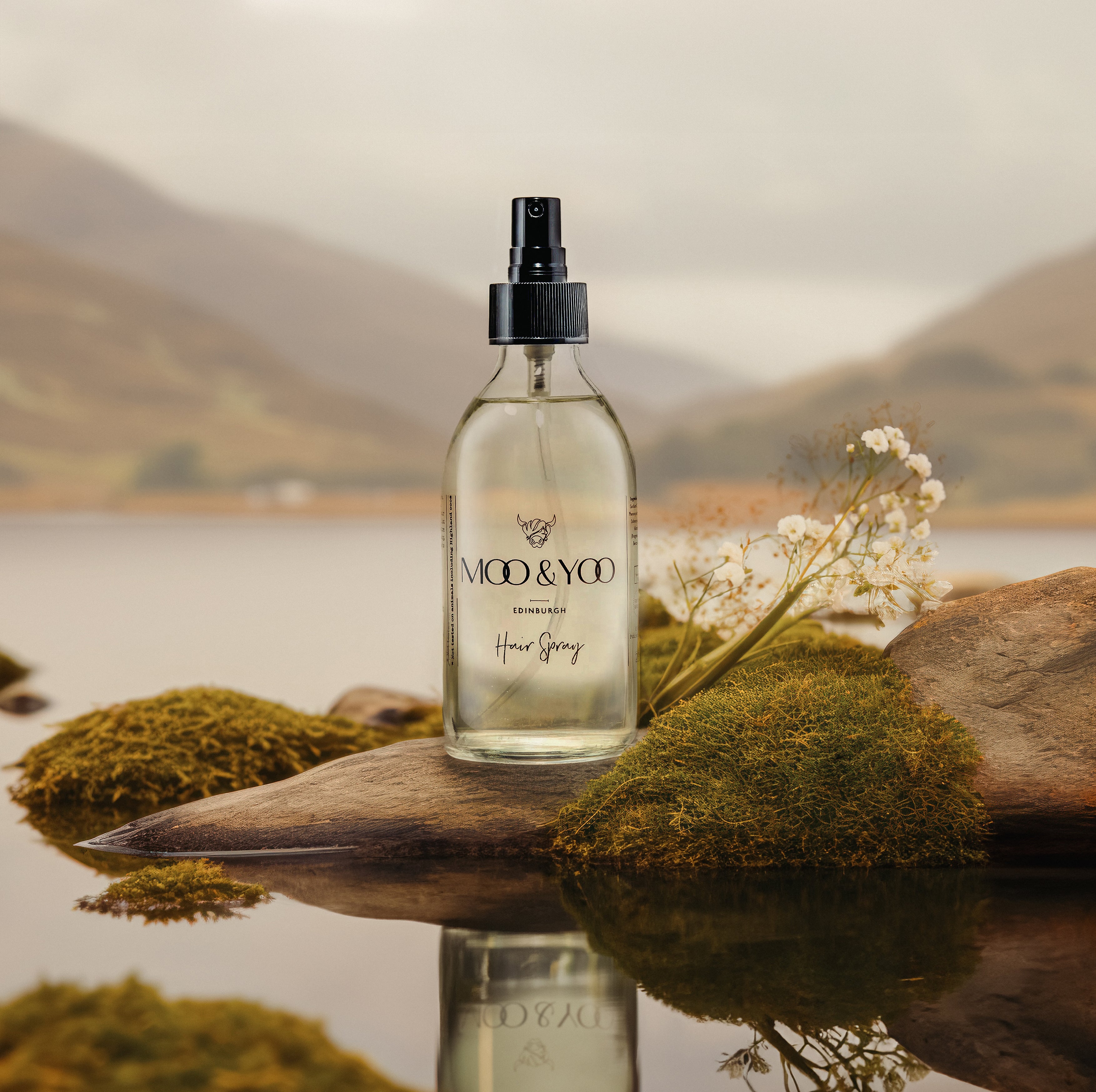 Our hair spray in a glass bottle on a small rock peaking out onto the loch with the water surrounding it and munros in the background 