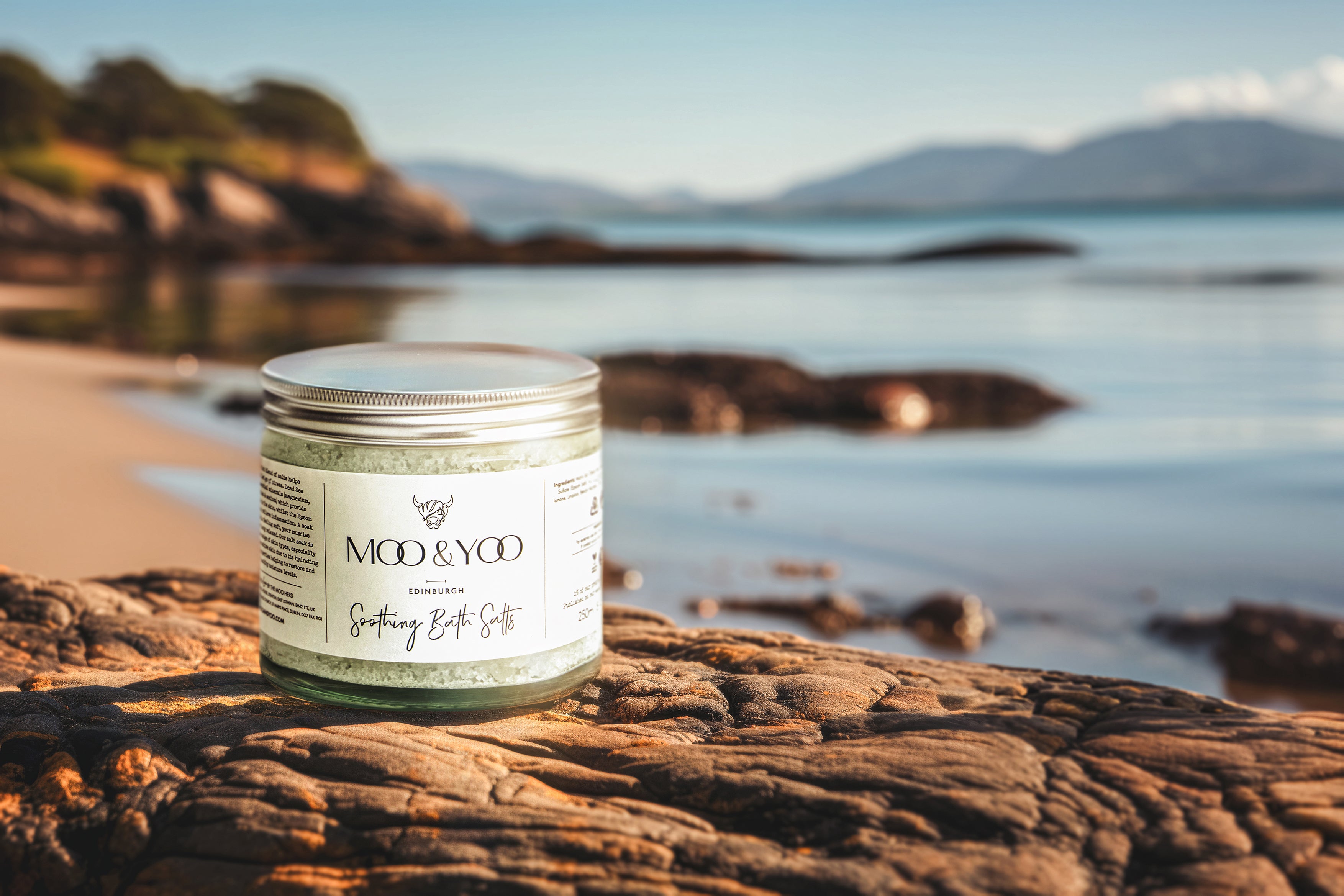 Our soothing bath salts in a glass jar on a rock in the foreground, a beach cove as the backdrop