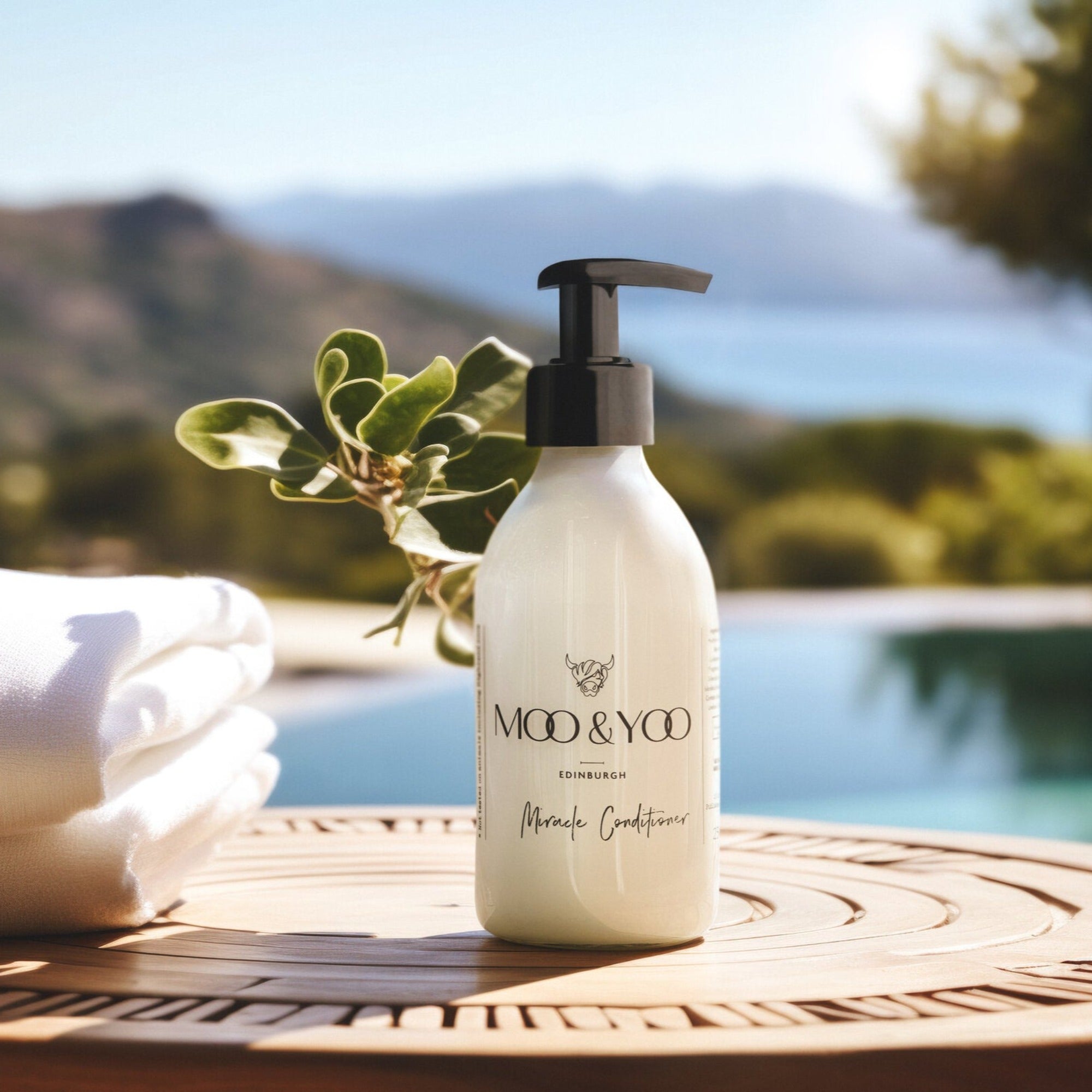 A close up image of a glass bottle of Moo and Yoo Miracle conditioner with a pump. It is placed on a table at a villa overlooking the swimming pool. There is a towel to the side. In the distance you can see hills and the sea