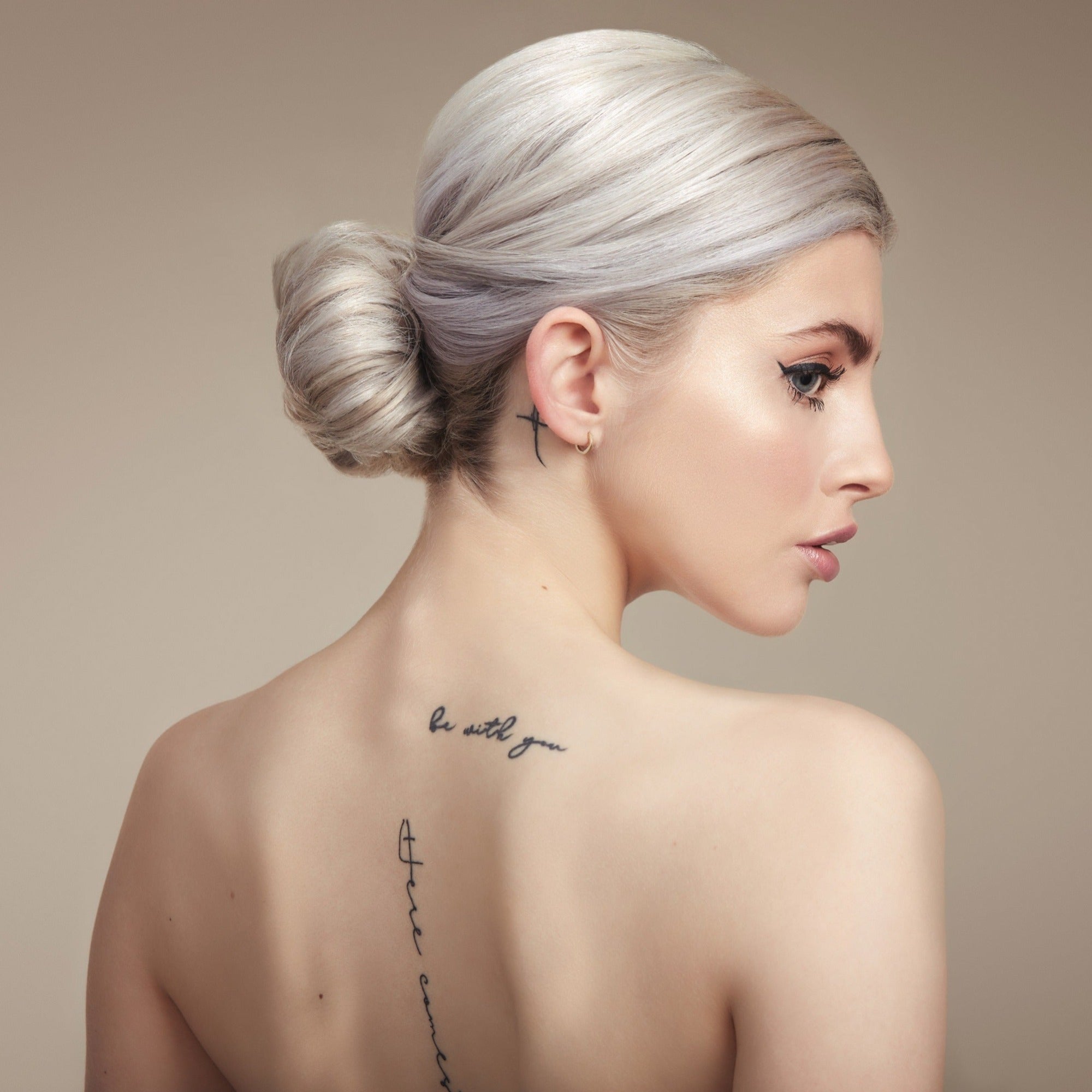 Female model with her back to the camera. Her platinum blonde hair is in a neat low bun, back is bare and she has tattoos down her spine