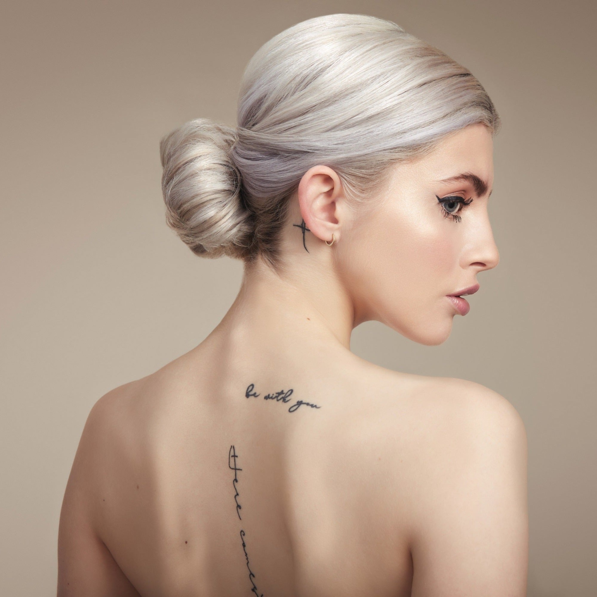 Female model with bleached blond hair in a low bun. She has her back to the camera so you can see the bun and she is turning her head to the side so you can see her profile. She has processed hair so has used Moo and Yoo Miracle Conditioner and Mask. Her back is bare and she has some tattoos.