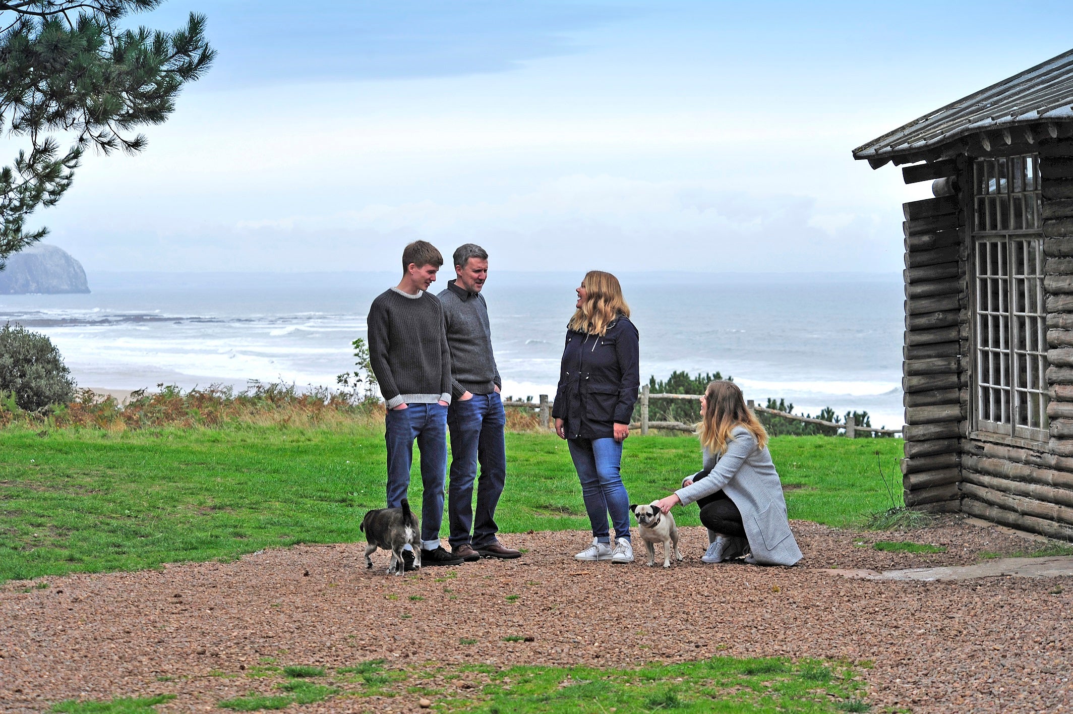the moo and yoo fam,ily on the scottish east coast with the dogs. There is a wooden cabin on the right, the beach in the background and they are standing on the hill, talking and laughing together. The family consists of Ali (left), Scott (middle left), Suzie (middle right) and Olivia (far right).