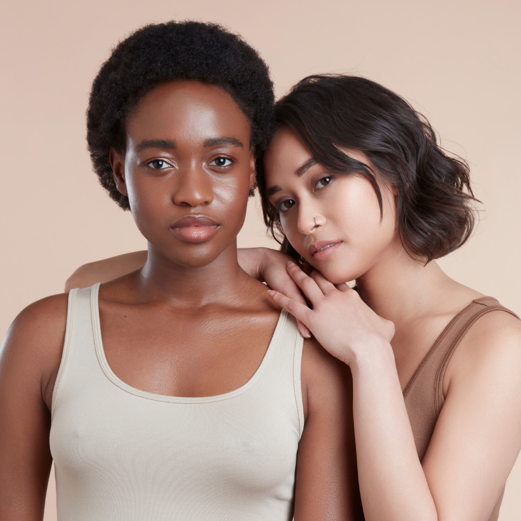 Image of twofemale models looking at the camera. One has bob hair, the other one has black textured natural hair. They are wearing neutral vest tops. Their skin is glowing and thier hair is healthy and shiny