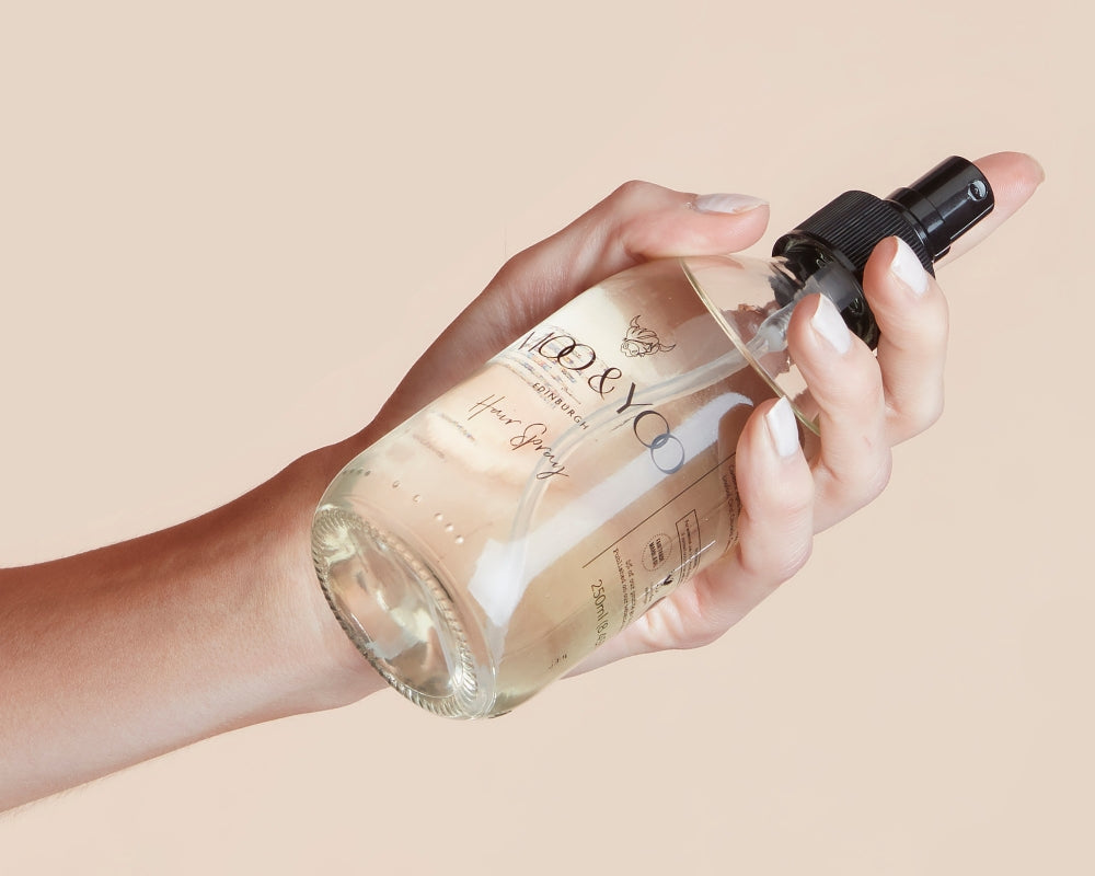 Hand holding a glass bottle of moo & yoo aerosol free hair spray on a neutral background