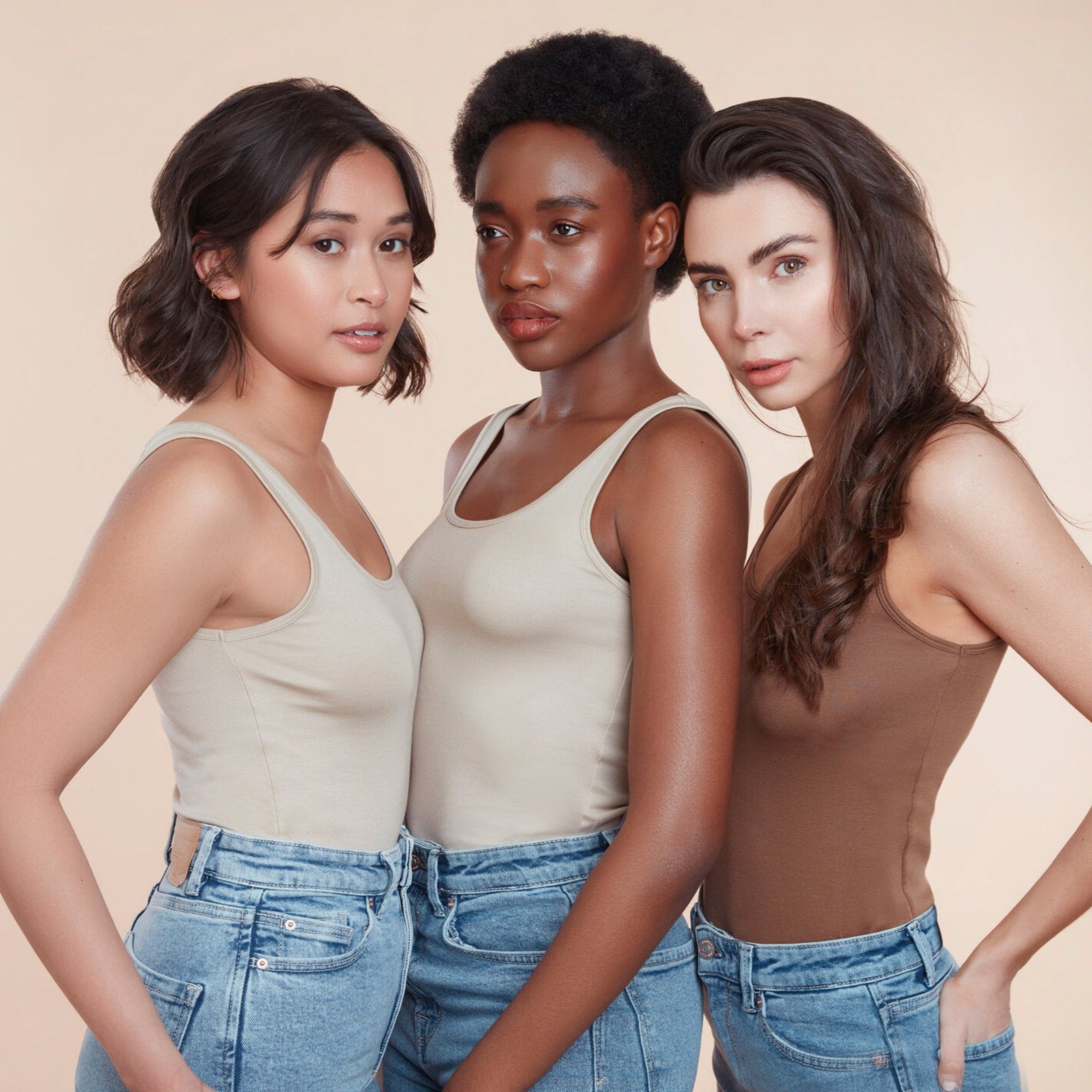 Three female models with moo and yoo products in their hair all wearing jeans and neutral coloured vest tops. They are looking directly at the camera. All have glowing skin and healthy shiny hair. One girl has bob length wavy hair, One girl has black natural textured hair and the other girl has long brown hair.