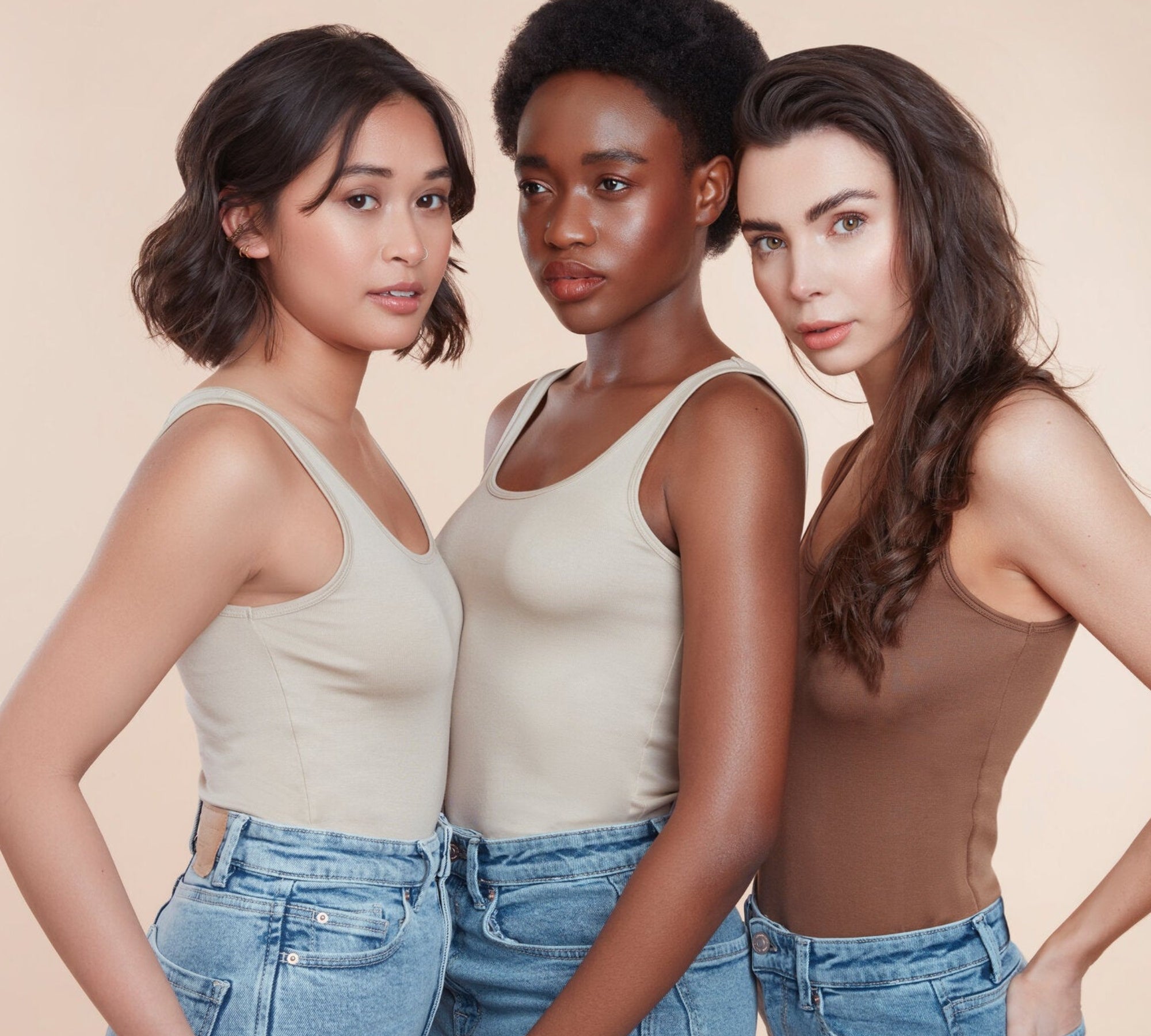 Three female models with moo and yoo products in their hair all wearing jeans and neutral coloured vest tops. They are looking directly at the camera. All have glowing skin and healthy shiny hair. One girl has bob length wavy hair, One girl has black natural textured hair and the other girl has long brown hair. 
