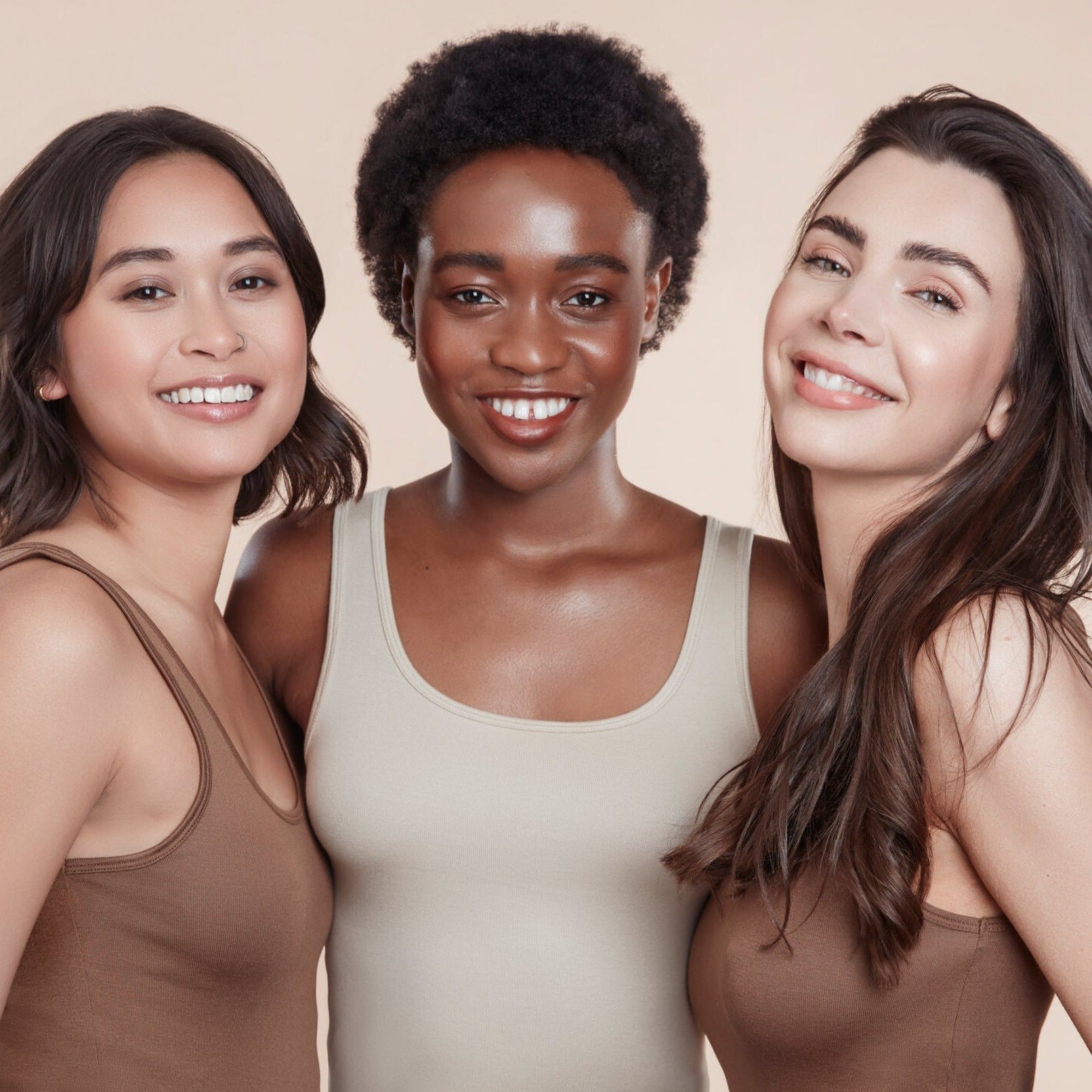 Three female models with moo and yoo products in their hair all wearing neutral coloured vest tops. They are looking directly at the camera smiling. All have glowing skin and healthy shiny hair. One has bob length wavy hair, One has black natural textured hair and one has long brown hair.