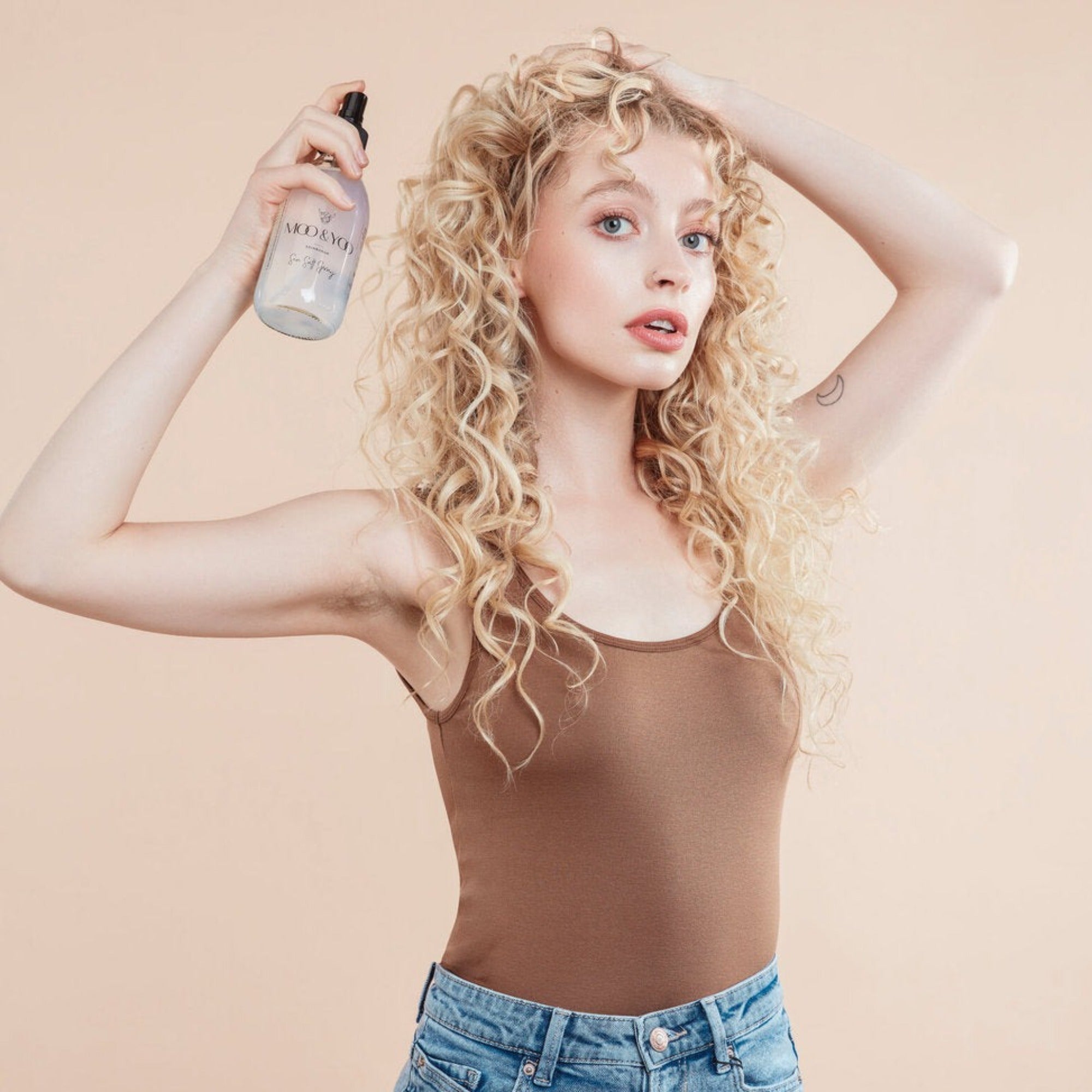Female model with flowing blond curly hair. She is holding Moo and Yoo Sea Salt Spray in her right hand and spraying it into her hair, she is touching her hair with her left hand. She is wearing a simple natural coloured vest top and her make up is very natural.