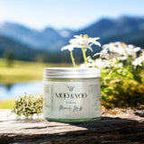 A glass jar of Moo and Yoo Miracle Mask with an aluminium lid. It is placed on a rustic wooden bench overlooking a Scottish Loch and hills. It is a spring day and there are white spring flowers behind it.