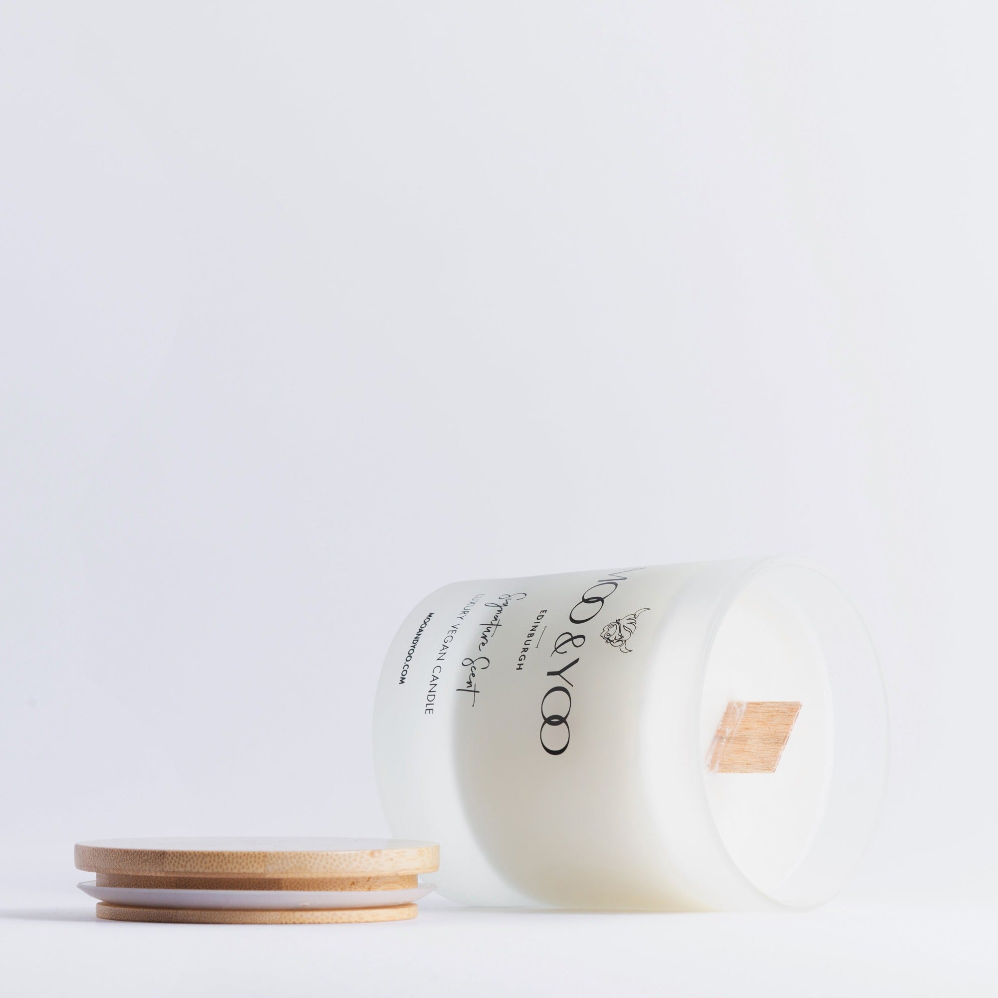 Glass jar with bamboo lid of Moo and Yoo signature scent luxury vegan candle. It is on its side showing the wooden wick with the lid to the side and on a white backgroun d.