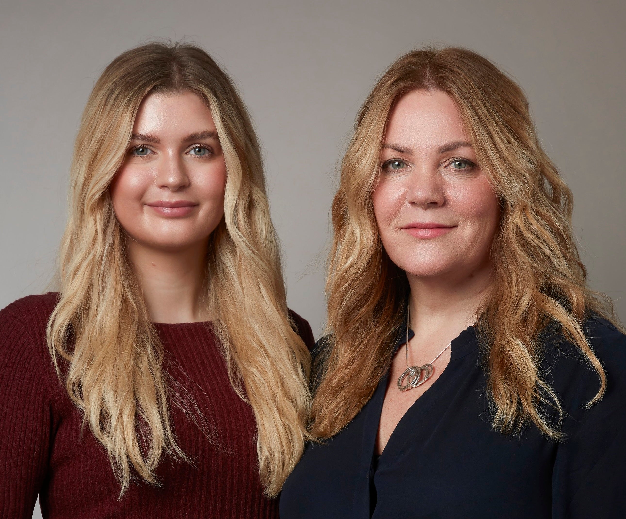 Studio shot of mother and daughter Suzie and Olivia. Both have long blond hair in loose waves, smiling at the camera 