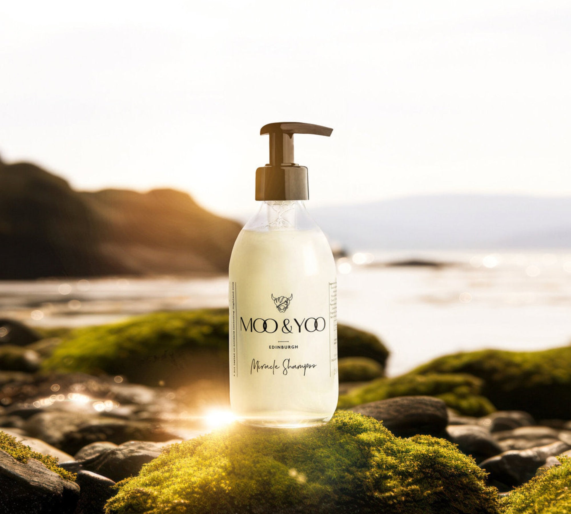 A glass bottle of Moo and Yoo Miracle shampoo with a pump on the edge of a Scottish Loch. The sun is reflecting off the bottom of the glass bottle creating a glow.