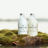 A glass bottle of Moo and Yoo Miracle Conditioner and a glass bottle of Moo and Yoo Miracle Shampoo sitting side by side with aluminium lids. They are placed on a rock with moss in the edge of the water.