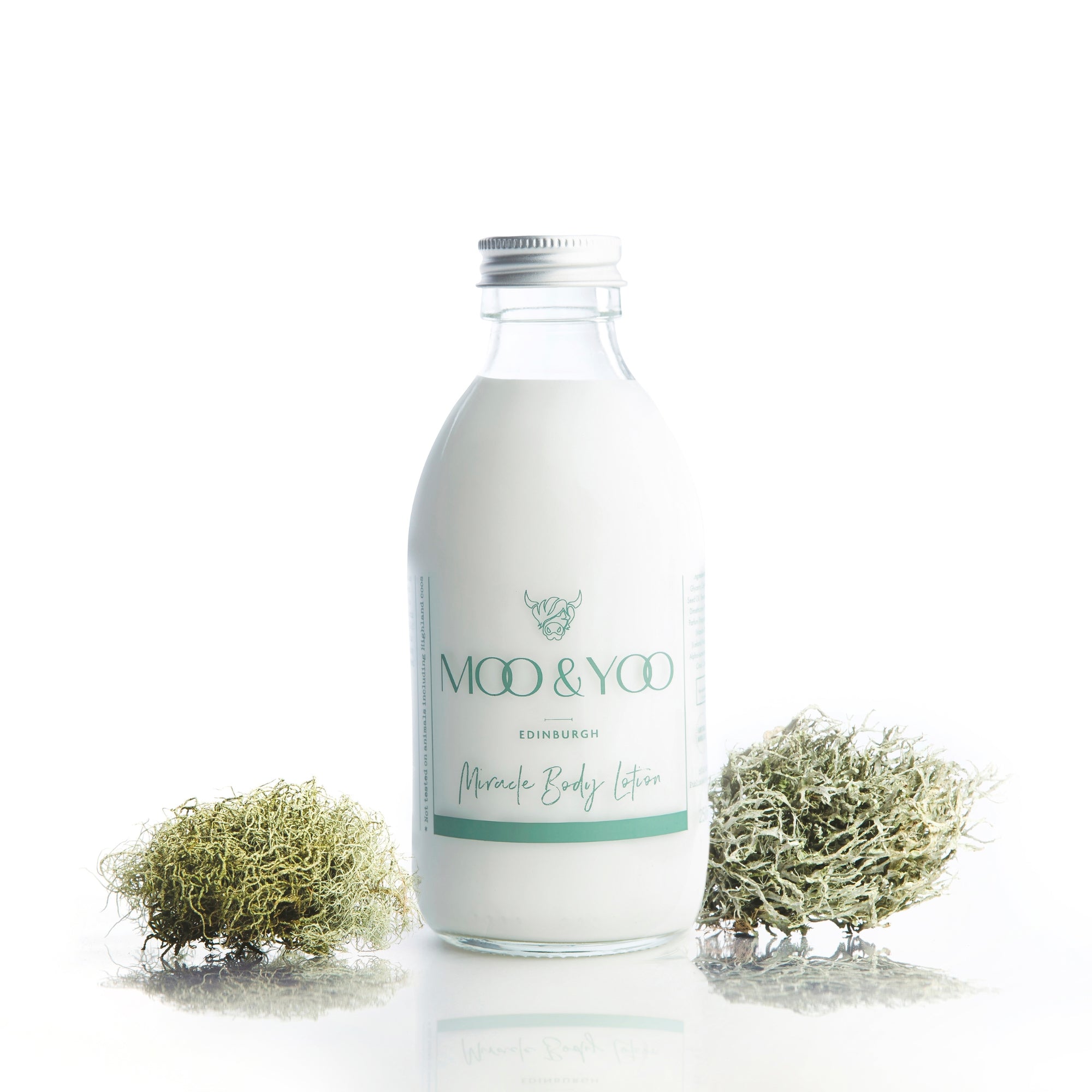 studio shot of our miracle body lotion with an aluminium lid in a glass bottle. There is green sprigs of moss on either side against a white