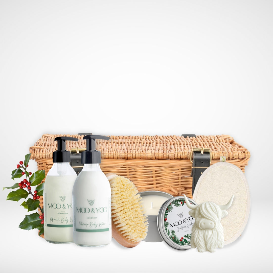 Luxury body care product hamper with our vegan body wash and body lotion in glass bottles, a sisal dry body brush, christmas scented tin vegan candle, body buffer pad and a highland cow shaped hand soap all infront of a wicker hamper basket with buckles against a white background