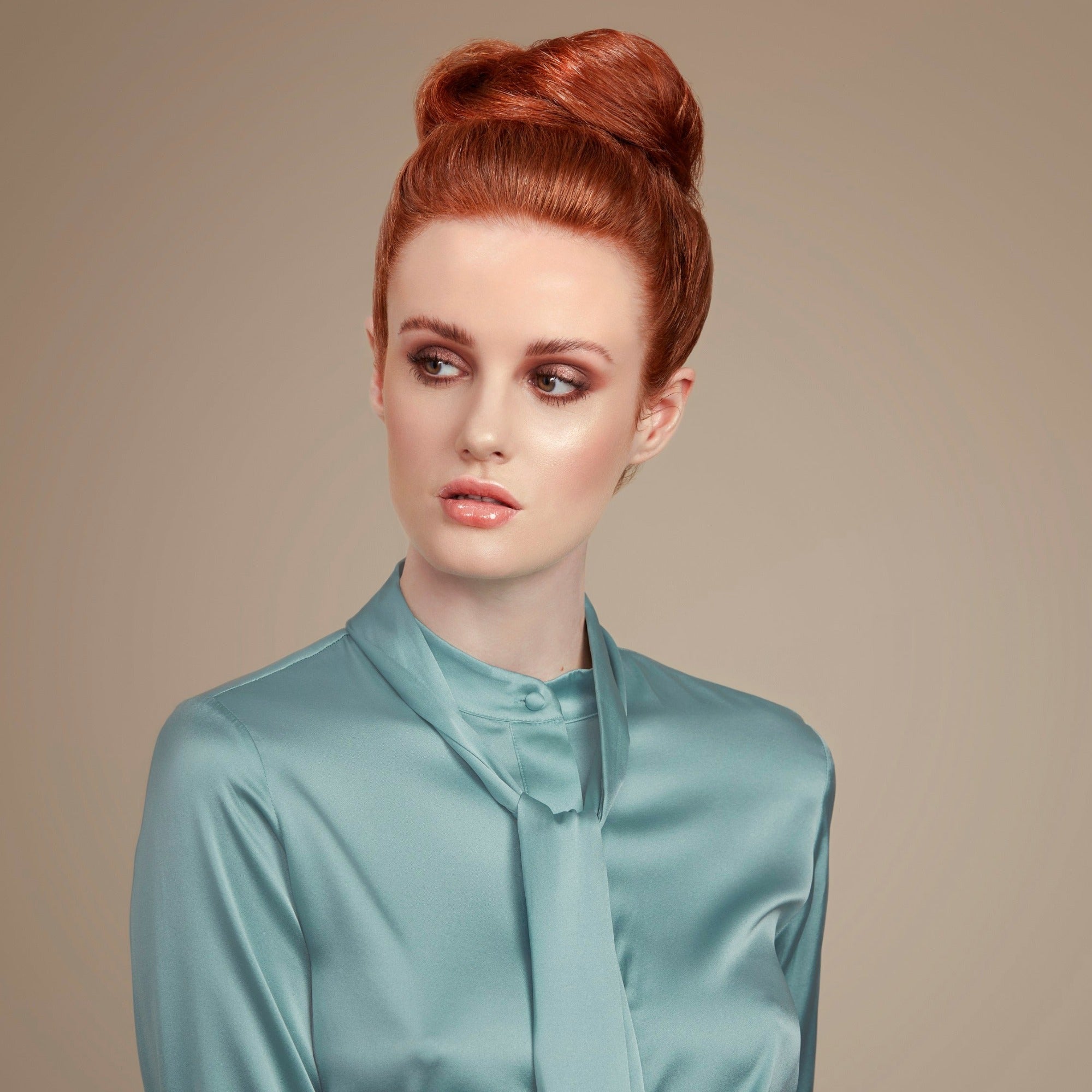 Female model with red hair in a high bun held in place with Moo and Yoo hair spray. She is facing the camera and looking to her left. She is wearing a pale blue top that is long sleeved and has a high neck.
