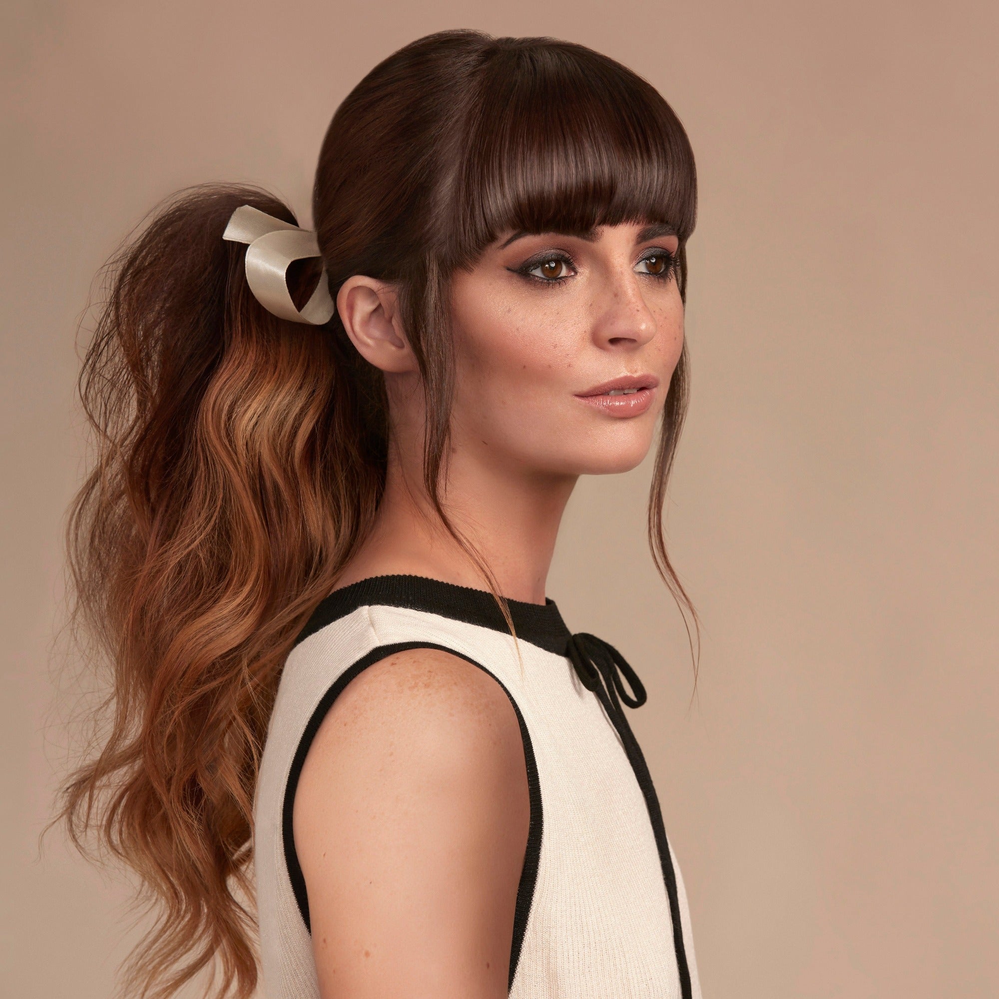 Female model with long wavy brunette hair tied into a low pony tail with a cream ribbon and a straight fringe framing her face. Her hair is very shiny from using Moo and Yoo Miracle Conditioner and full of volume form Moo and Yoo Volumising Spray Mist with a touch of Sea Salt Spray for texture. She is wearing a simple white dress with black trim.
