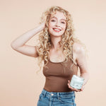 Woman with curly blonde hair holding a jar of miracle mask while feeling her hair against a beige background