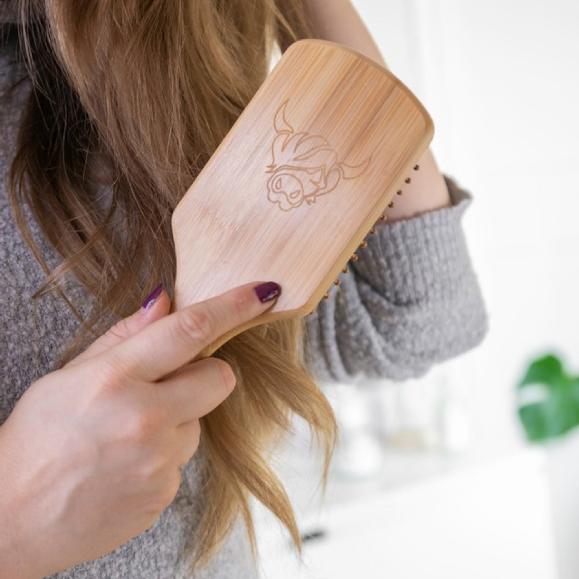 Person brushing their blond hair with wooden hairbrush with Moo & Yoo cow logo, wearing grey jumper