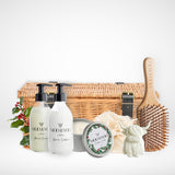 Hamper of hair care products including glass shampoo and conditioner bottles, a vegan christmas candle, a bamboo hair brush, and a small cow shaped hand soap, a fabric eco net shower ramie and a wicker hamper with black straps and buckles on a white background.