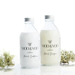 A glass bottle of Moo and Yoo Miracle Conditioner and a glass bottle of Moo and Yoo Miracle Shampoo sitting side by side with aluminium lids on a white background with a sprig of Icelandic moss placed to each side.