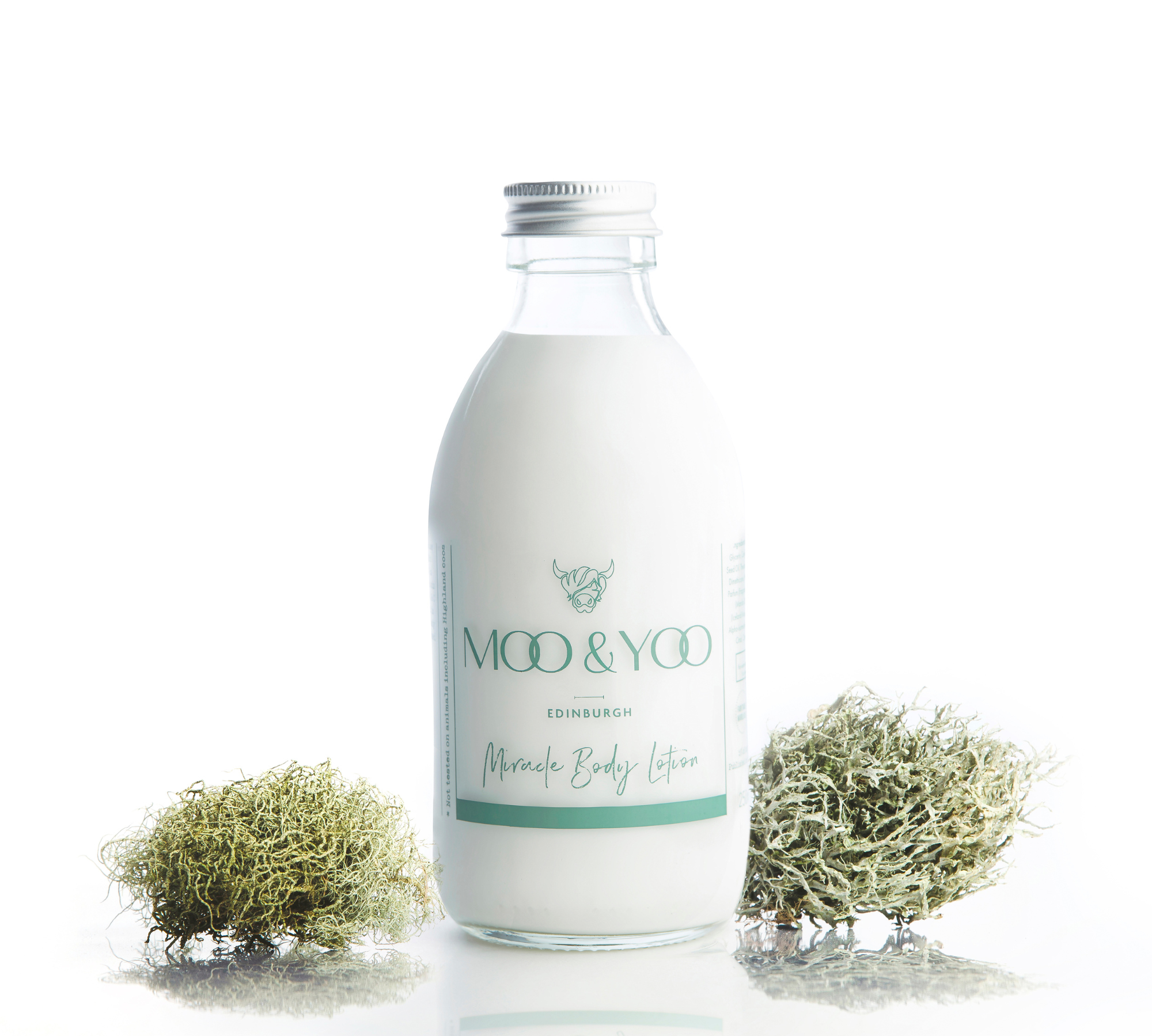 studio shot of our miracle body lotion with an aluminium lid in a glass bottle. There is green sprigs of moss on either side against a white background