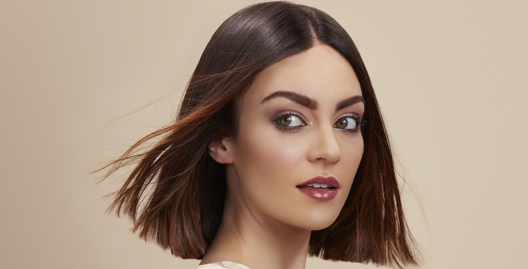 A female model with short, brunette, type 1A hair looking to her left with her hair being blown behind her. She is against a neutral, beige background.