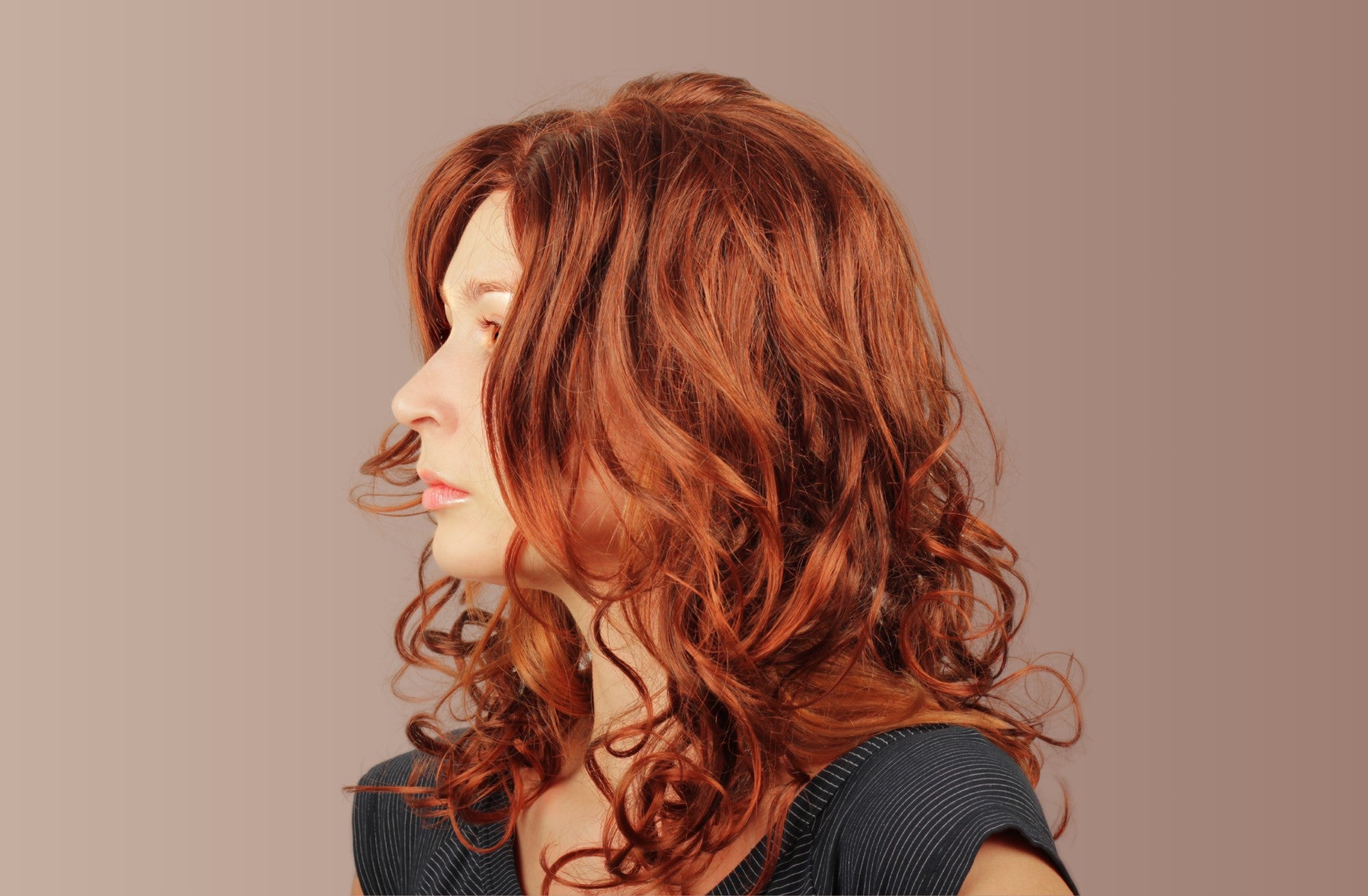 A female model with ginger, shoulder length, type 2A hair standing to the side with a neutral expression, against a brown background.