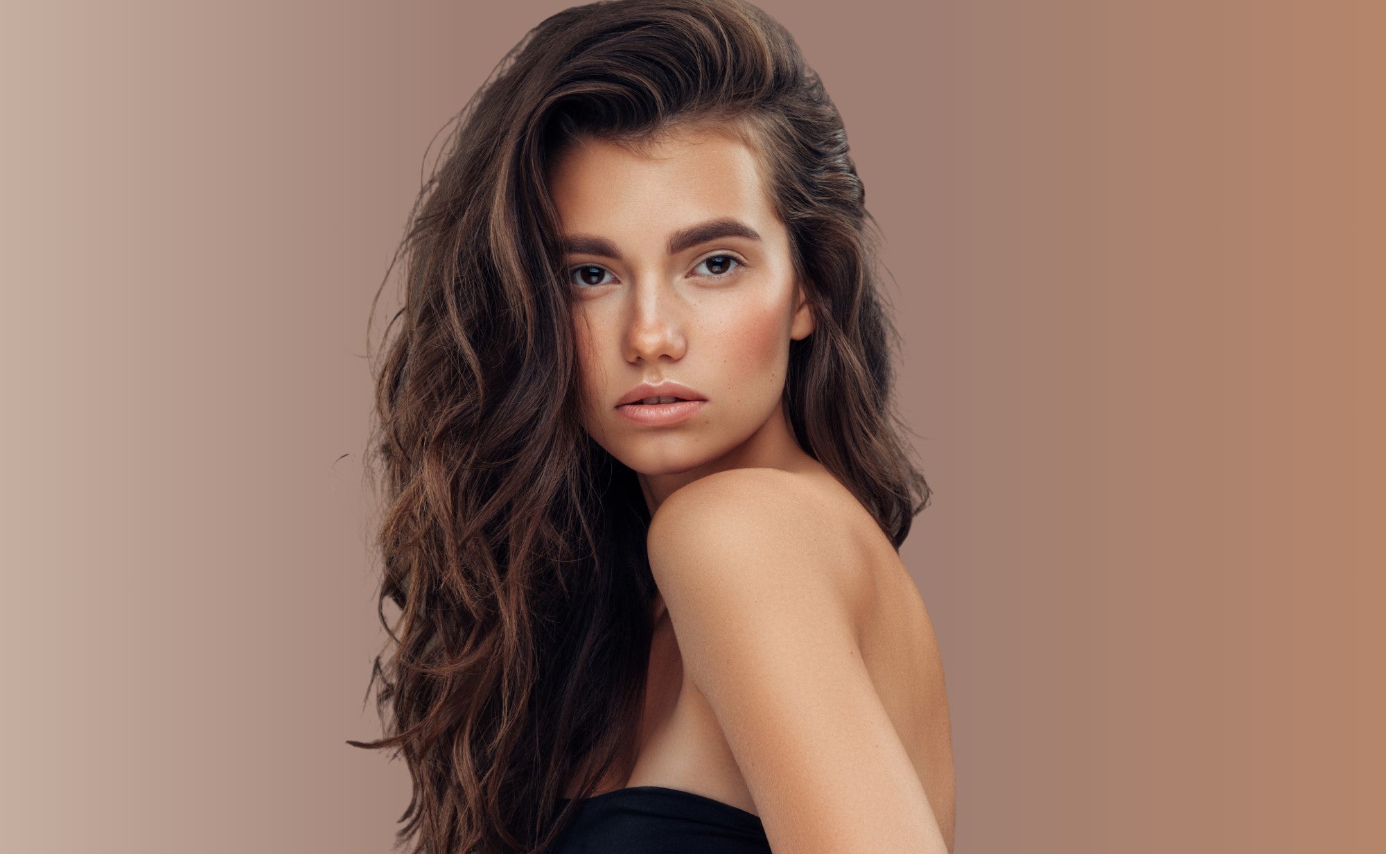 A female model with brown, wavy, type 2B hair looking over her shoulder at the camera with a neutral expression. She is against a beige/ brown background.