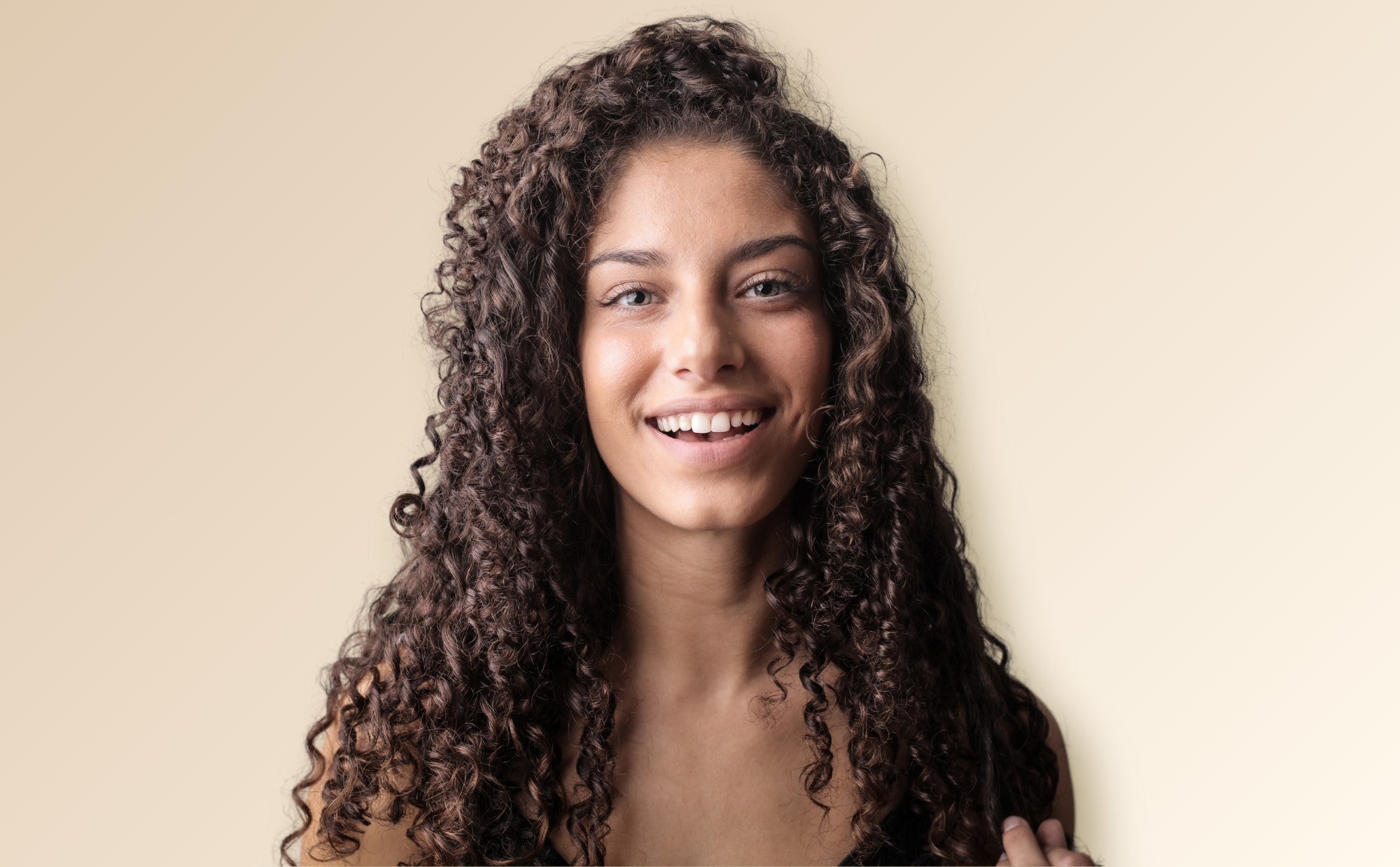 A female model with curly, dark brown, type 3B hair is in front of a neutral background and smiling at the camera.