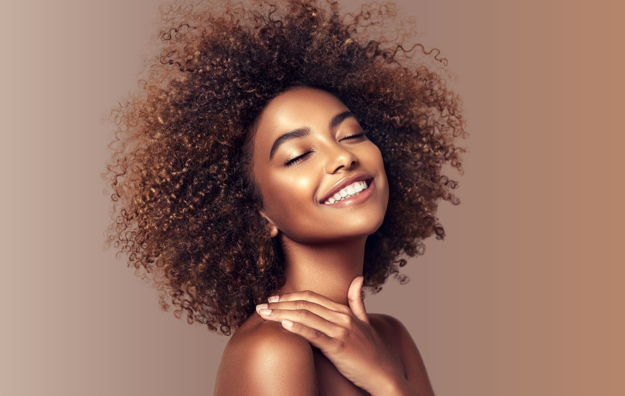 A female model with type 4A hair closing her eyes and smiling towards the camera, against a neutral, brown background.