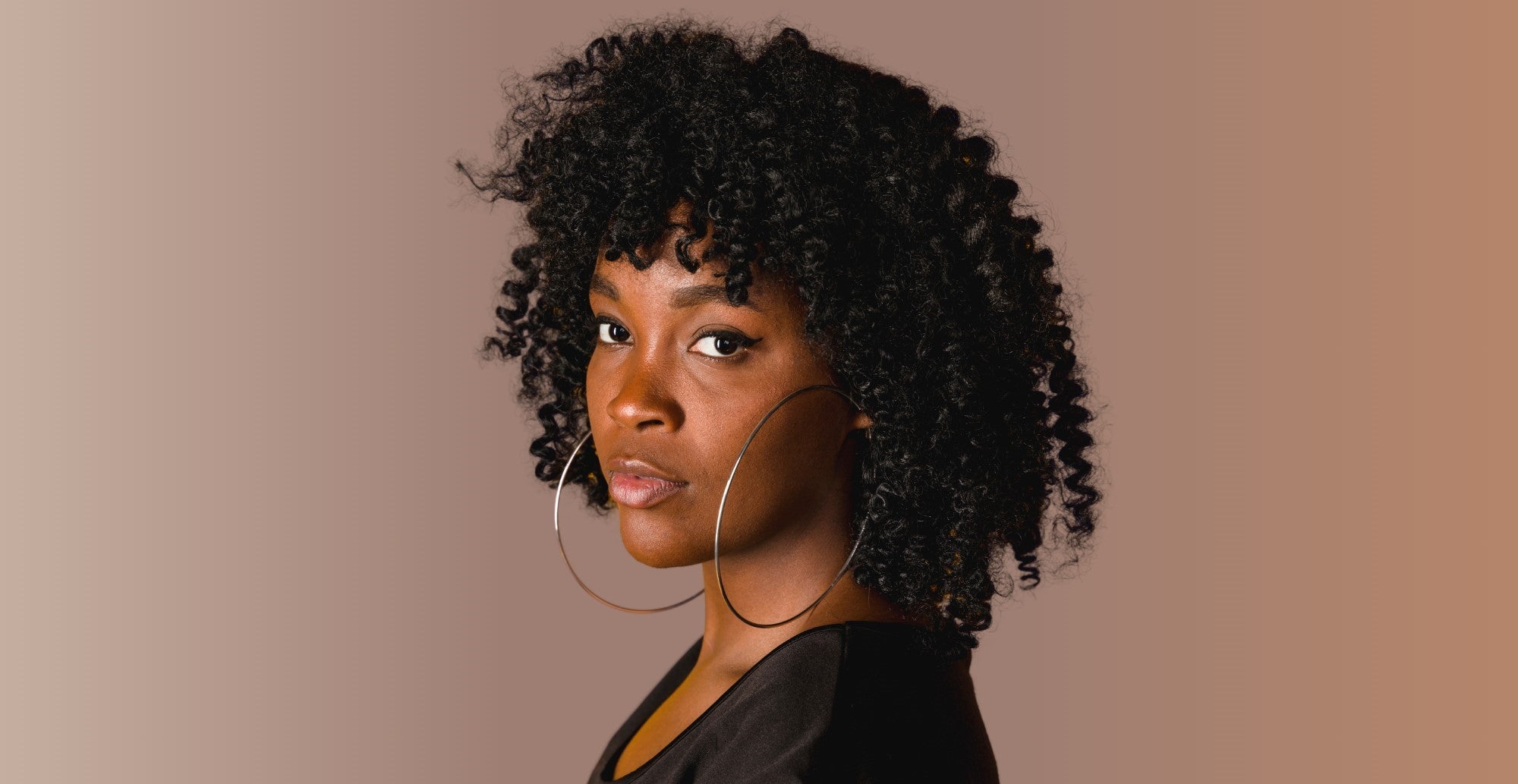 A female model with dark, type 4B hiar looking over her shoulder towards the camera with a neutral expression, against a brown background.