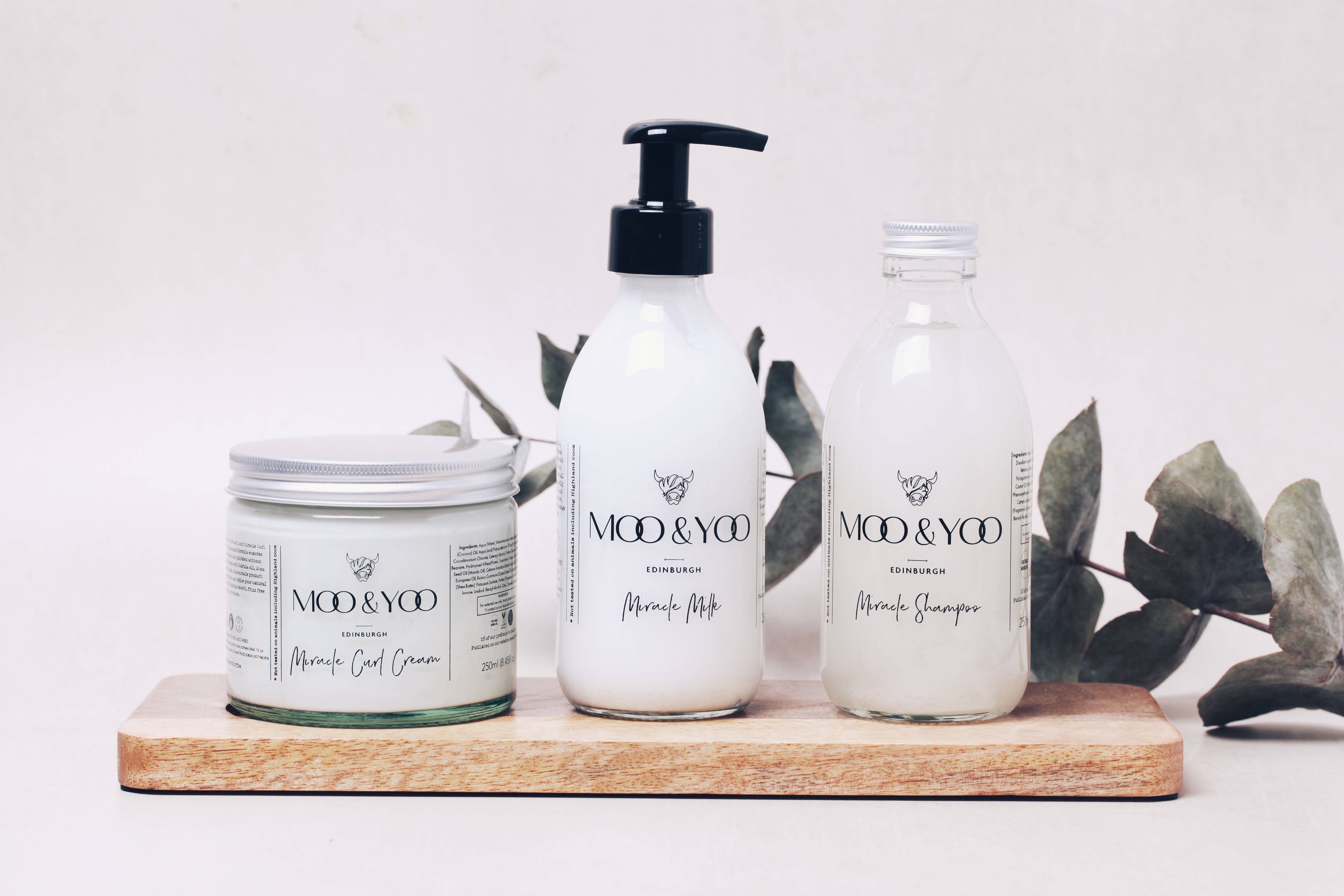 Three Moo & Yoo products, curl cream, miracle milk, and shampoo are lined up next to each other on a wooden baord. There is a plain backdrop behind them and a sprig of eucalyptus behind also.