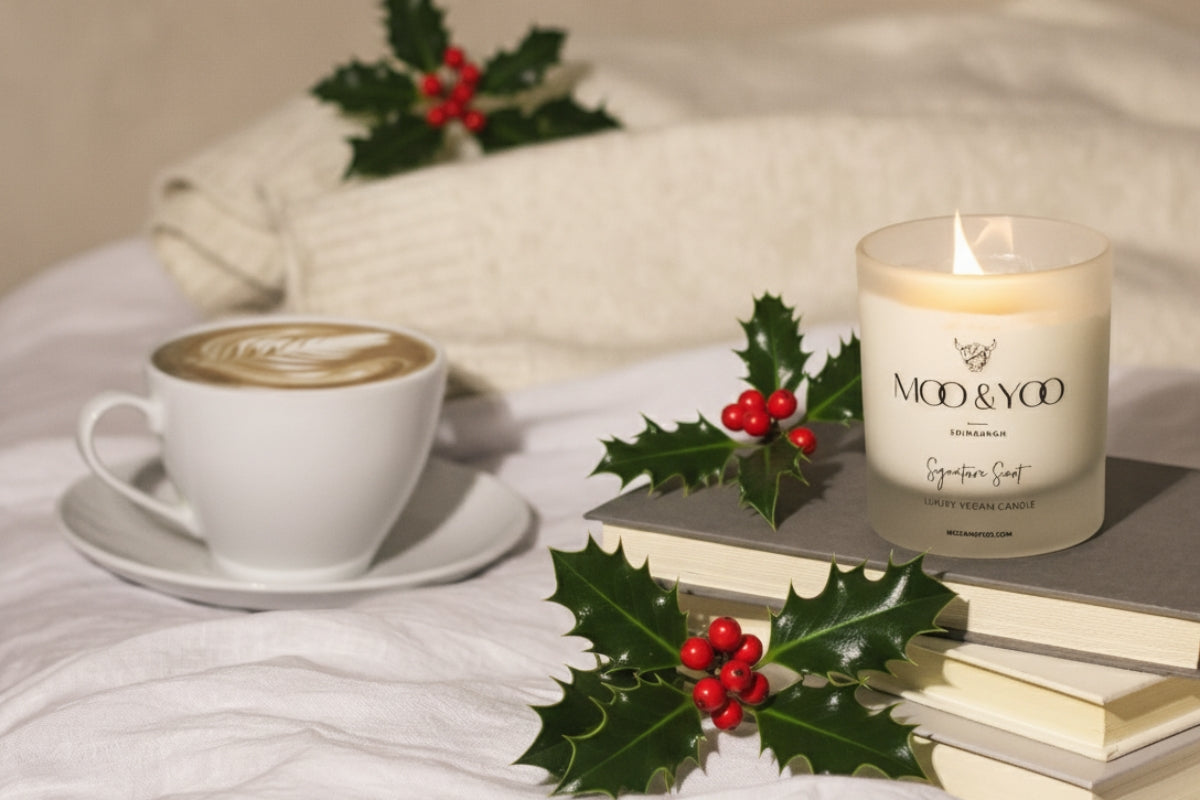 A cup of coffee, some books and a Moo & Yoo SIgnature Scent candle are placed on a bed along with some holly.