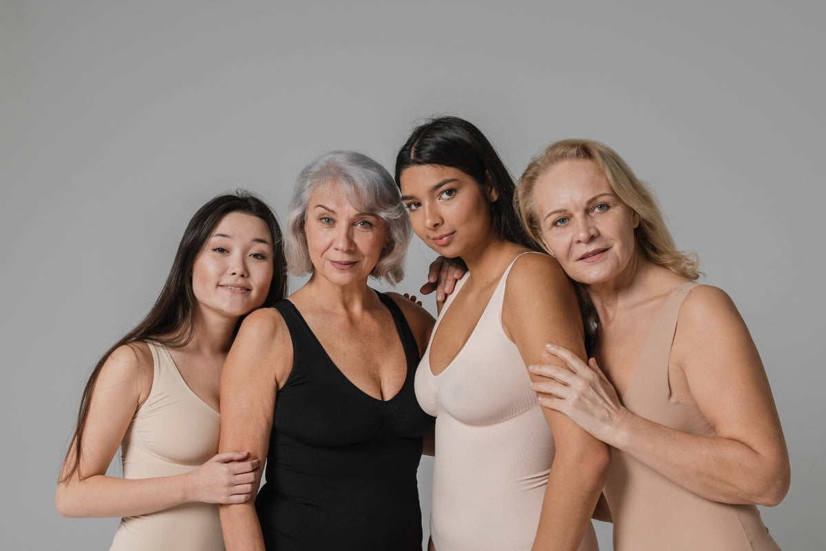4 women, 2 slightly older and 2 younger, are standing huddled togther in front of a neutral backgound. They are each wearing neutral tones and looking into the camera.