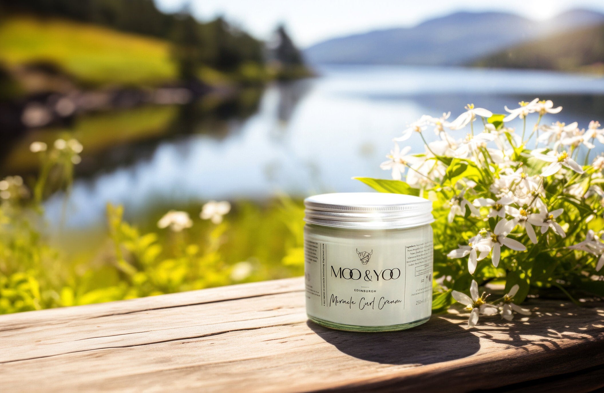 Jar of Moo & Yoo Miracle Curl Cream on a wooden bench in front of a scenic view of the sea, mountains and greenery