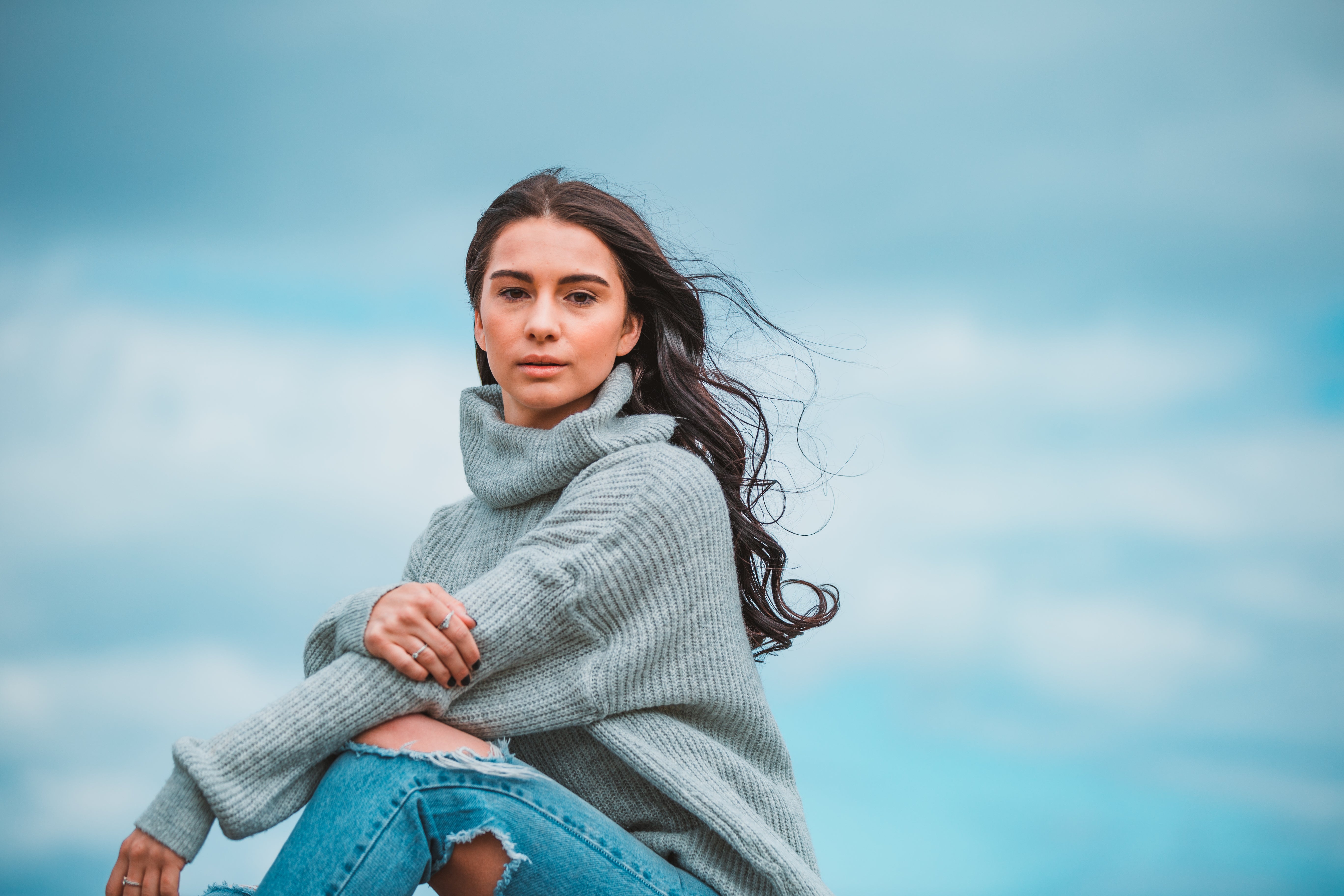 A female model with long, loose, curly hair is sitting with her knees up and her arms resting on her knees. She is looking at the camera with a neutral expression and there is a cloudy sky behind her.