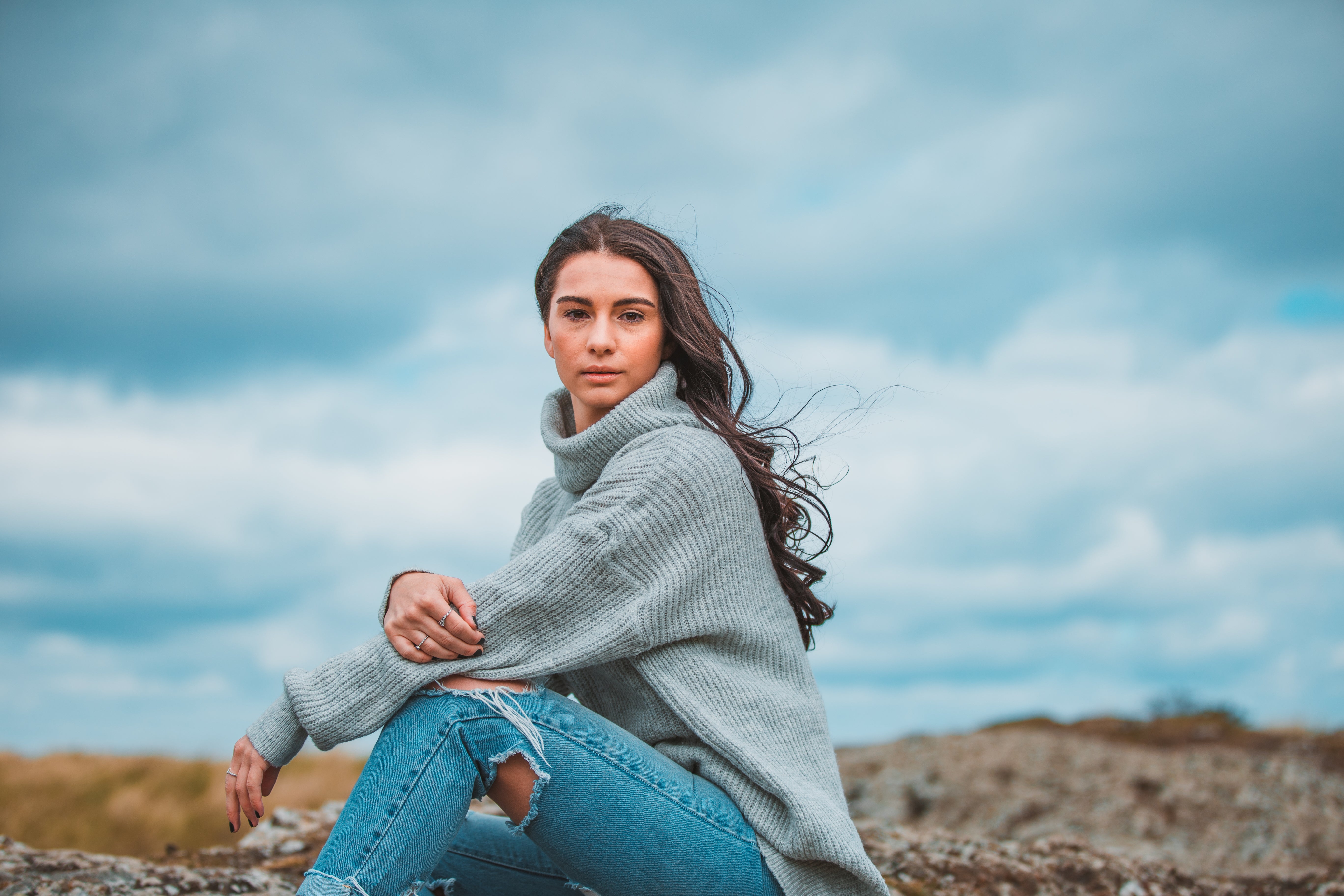 A model with long, wavy dark brown hair is sitting on a large rock looking at the camera. She has a neutral expression on her face. There is some dune grass in the background of the image and the sky is grey and cloudy.