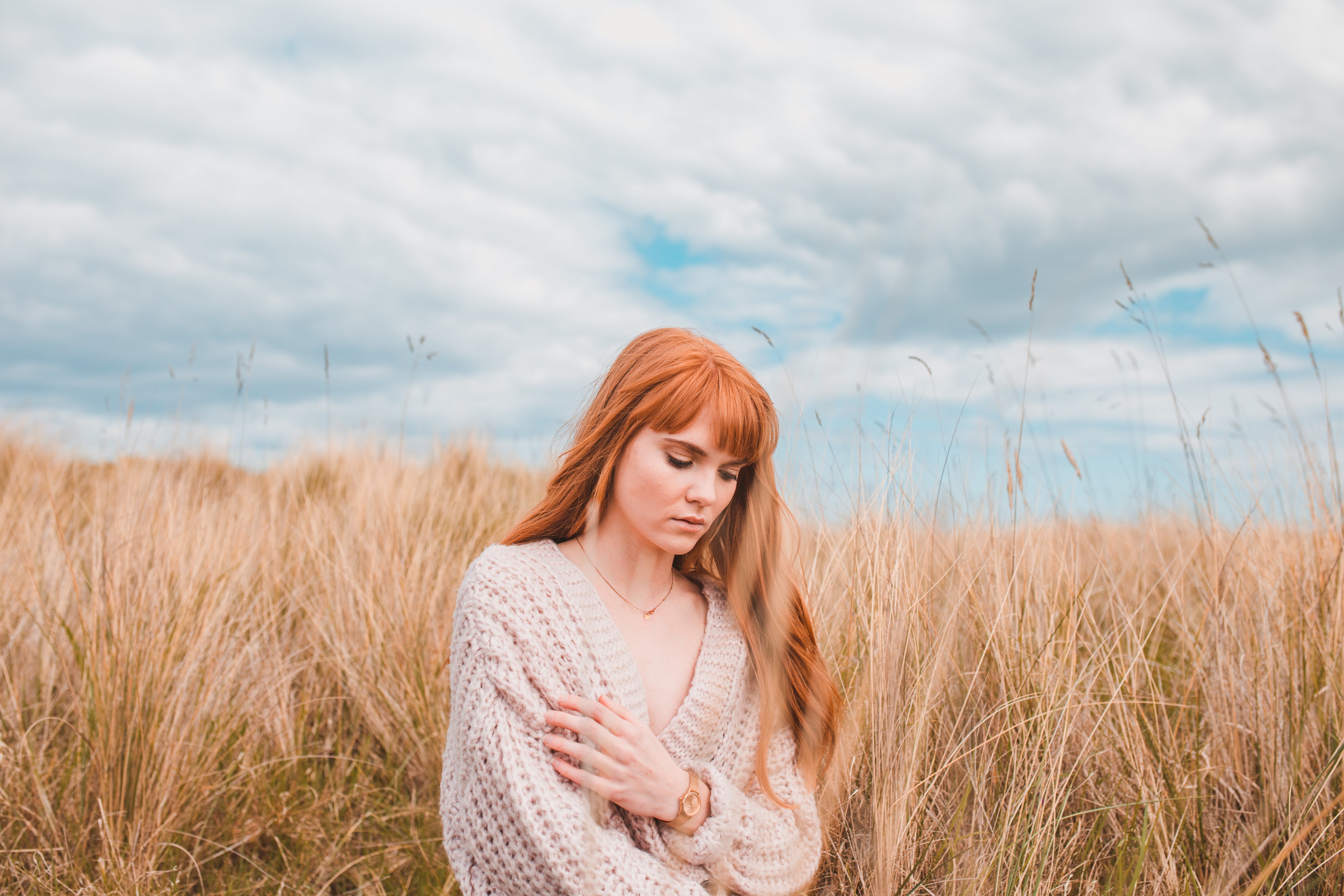 A female model with long flowing ginger hair is sitting in some dune grass. She has her arms wrapped around herself and is looking down towards the ground. She is wearing a knitted beige cardigan.