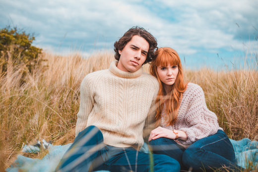 A man and women are sitting on a blanket in the middle of some dune grass. They are close together and more looking at the camera with neutral expressions. They are both wearing knitted jumpers.