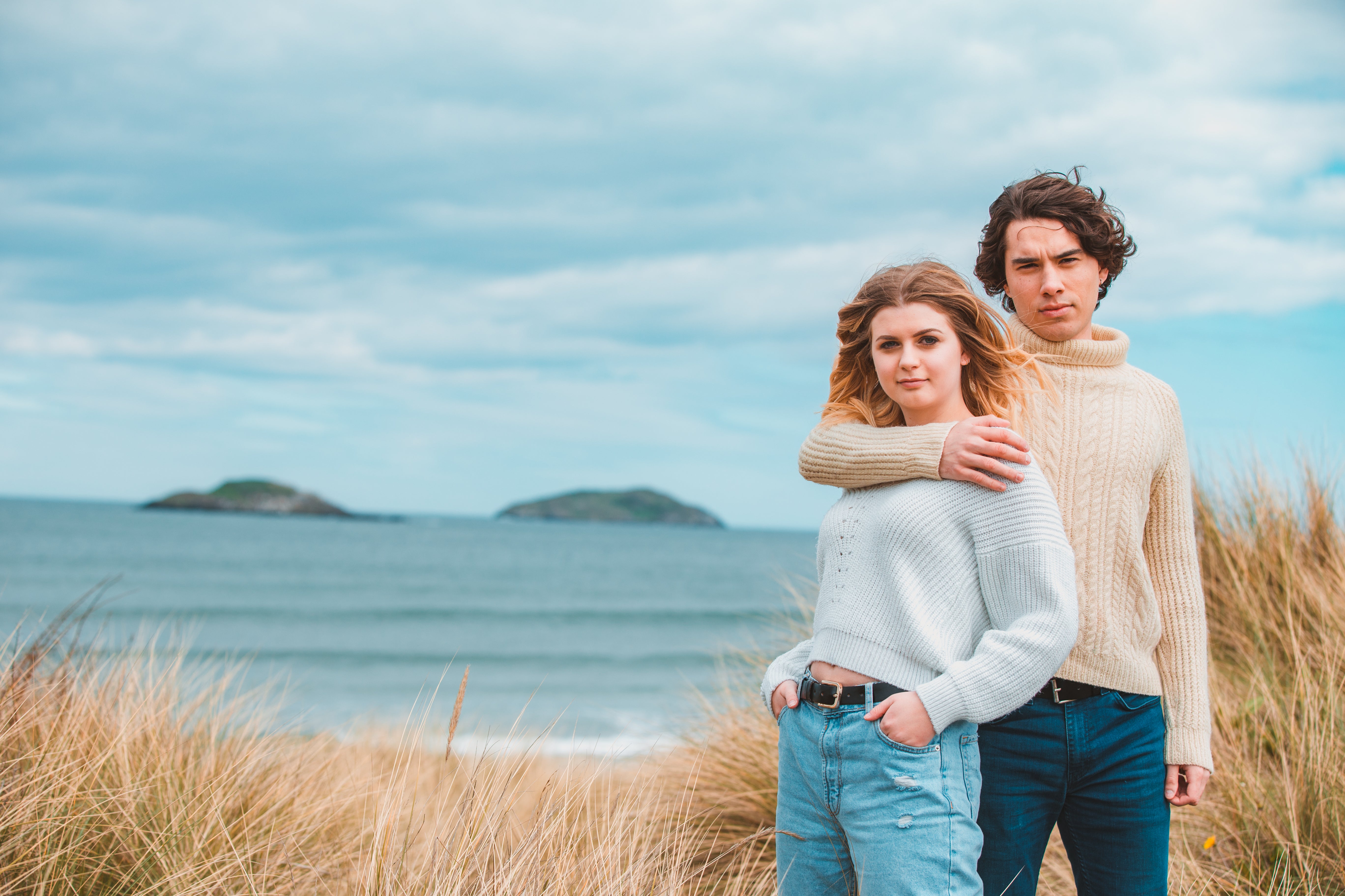 A man and woman are standing on the right of the image with the beach behind them. The man has one arm around the woman and they are both looking at the camera with a neutral expression.