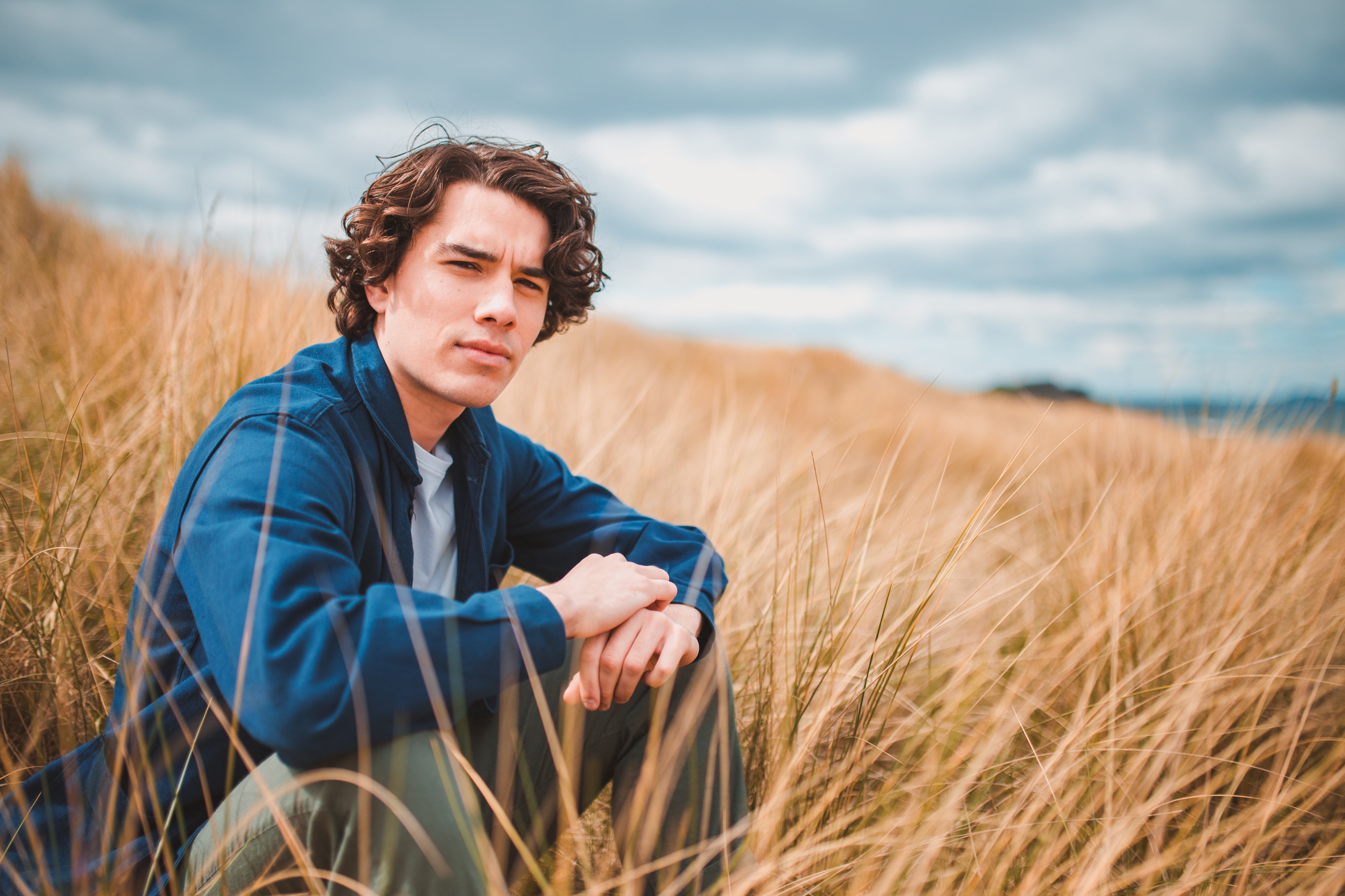 A male with short curly hair is sitting in some dune grass looking out towards the camera with a cloudy sky behind him. He has a neutral expression and his hair is flowing in the wind.