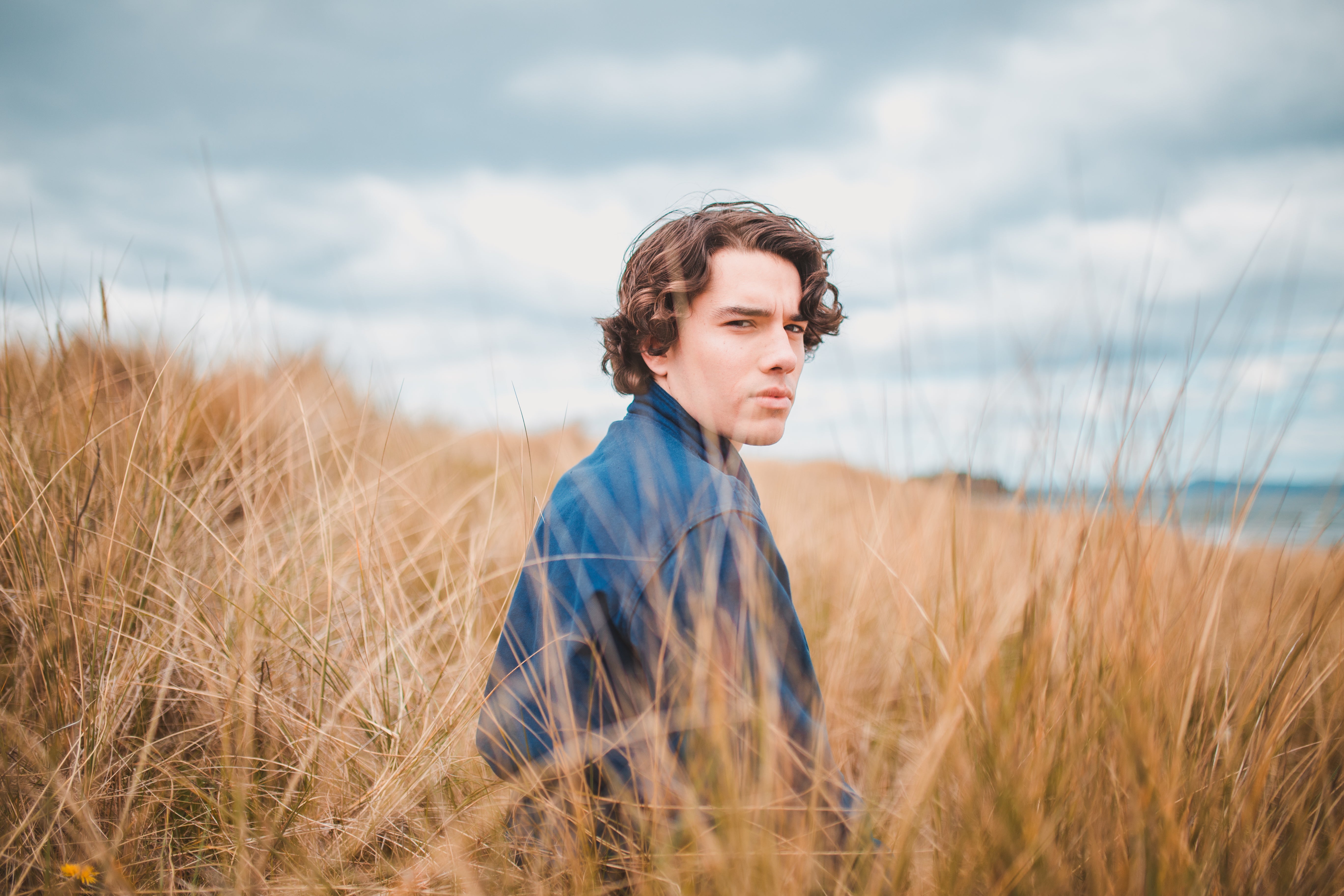 Male model is sitting in some dune grass. His body is facing away from the camera but his head is turned toward the camera. He has neutral facial expressions and has brown wavy hair.