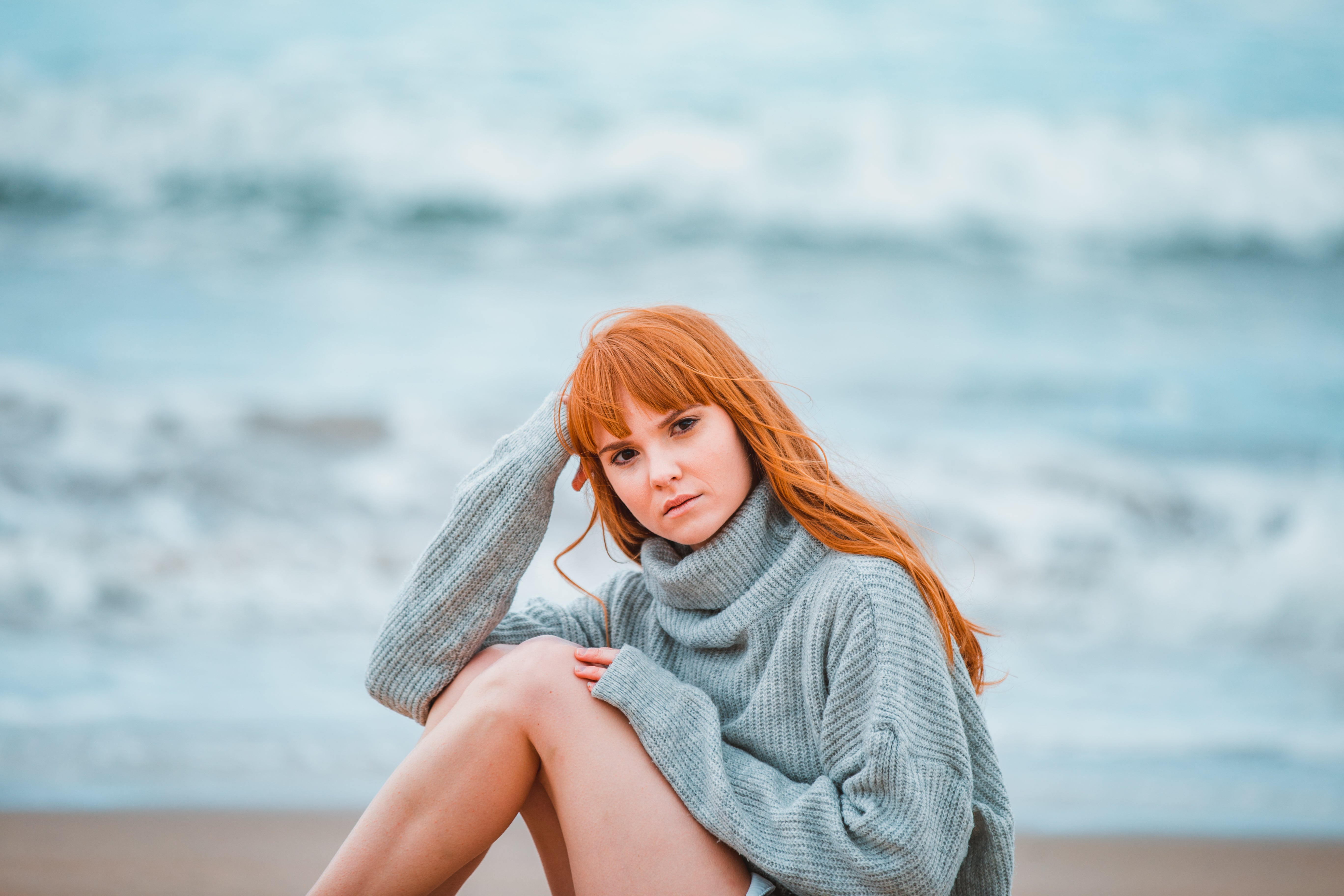 A female model with long flowing red hair is sitting on a beach. She is looking at the camera with a neutral expression. There is sand and waves in the background.