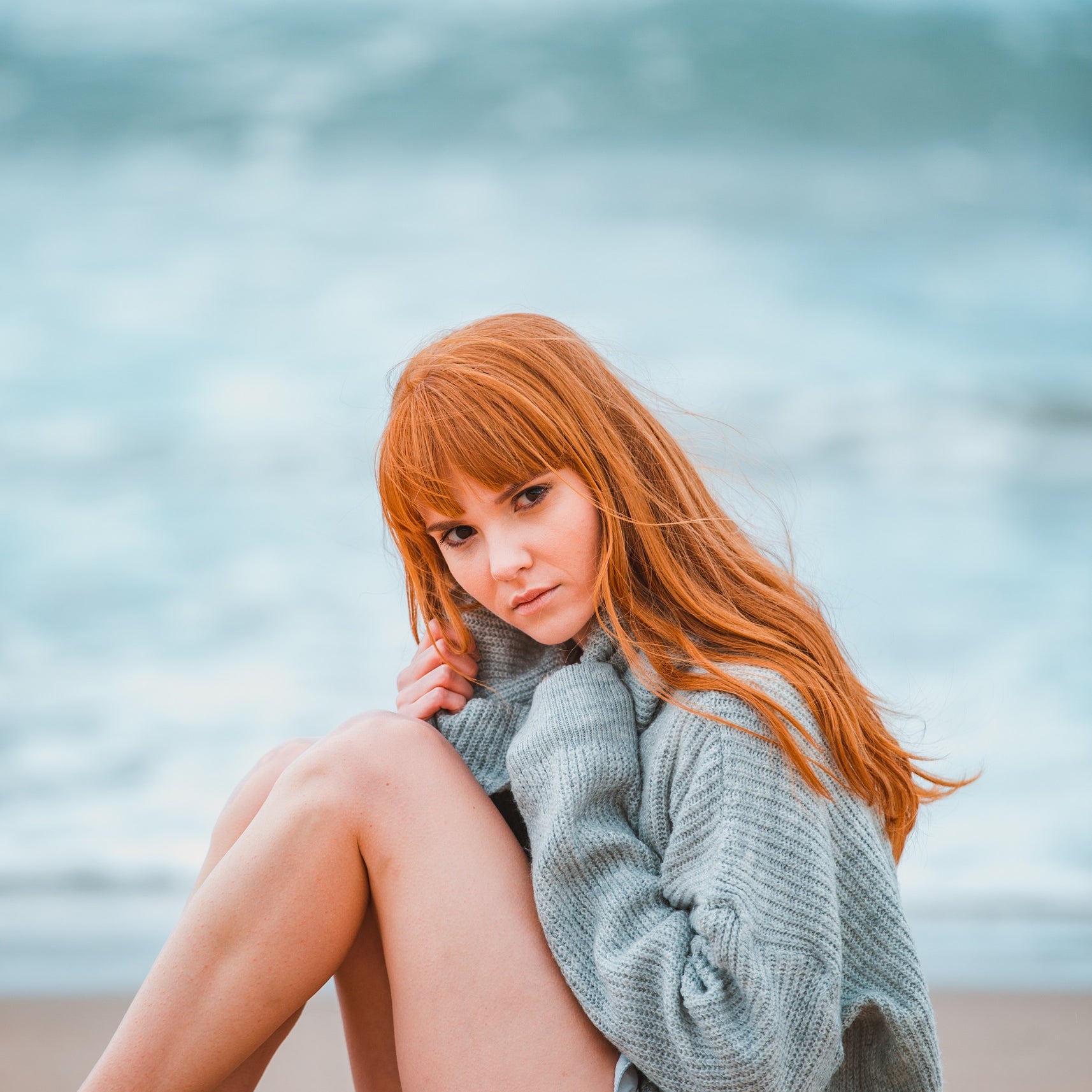 A female model is sitting on a beach with her knees drawn up to her chest. She has long, ginger, wavy hair with a fringe. She is looking at the camera with a neutral expression.