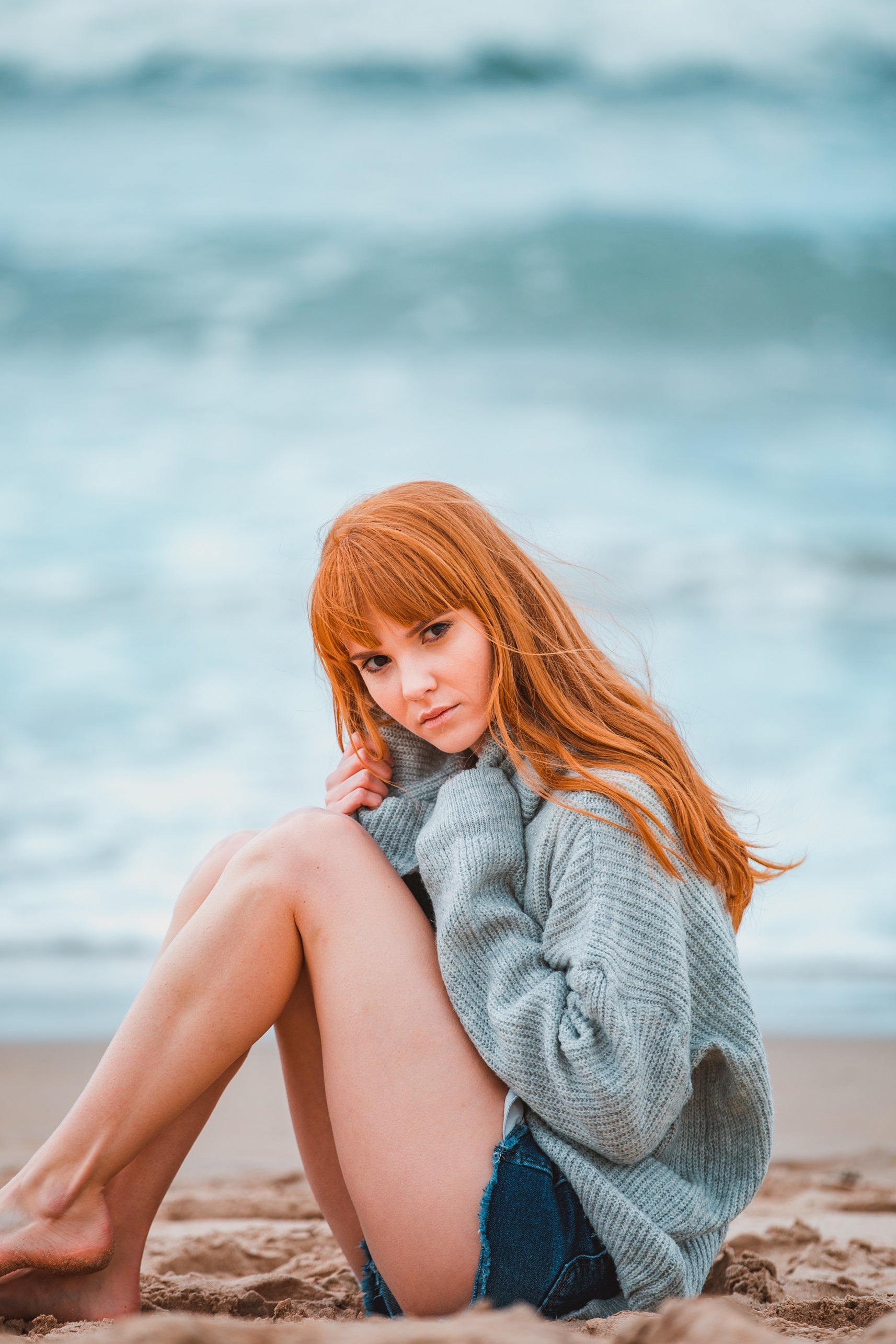 A female model is sitting on a beach with her knees drawn up to her chest. She has long, ginger, wavy hair with a fringe. She is looking at the camera with a neutral expression.