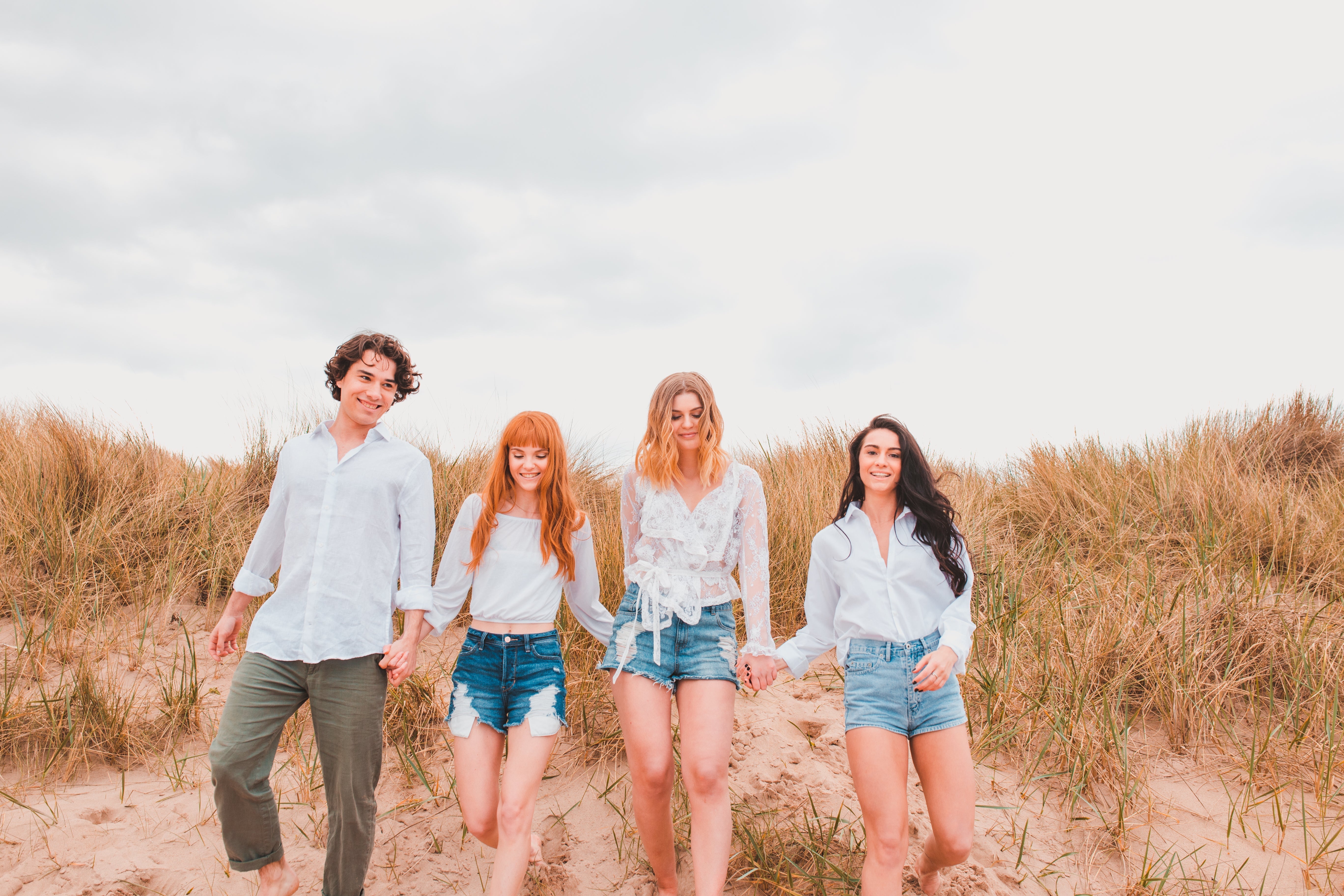 This image shows 4 people, 1 male and 3 females, walking down a sand dune all holding hands. They have dune grass behind them and they are all smiling.