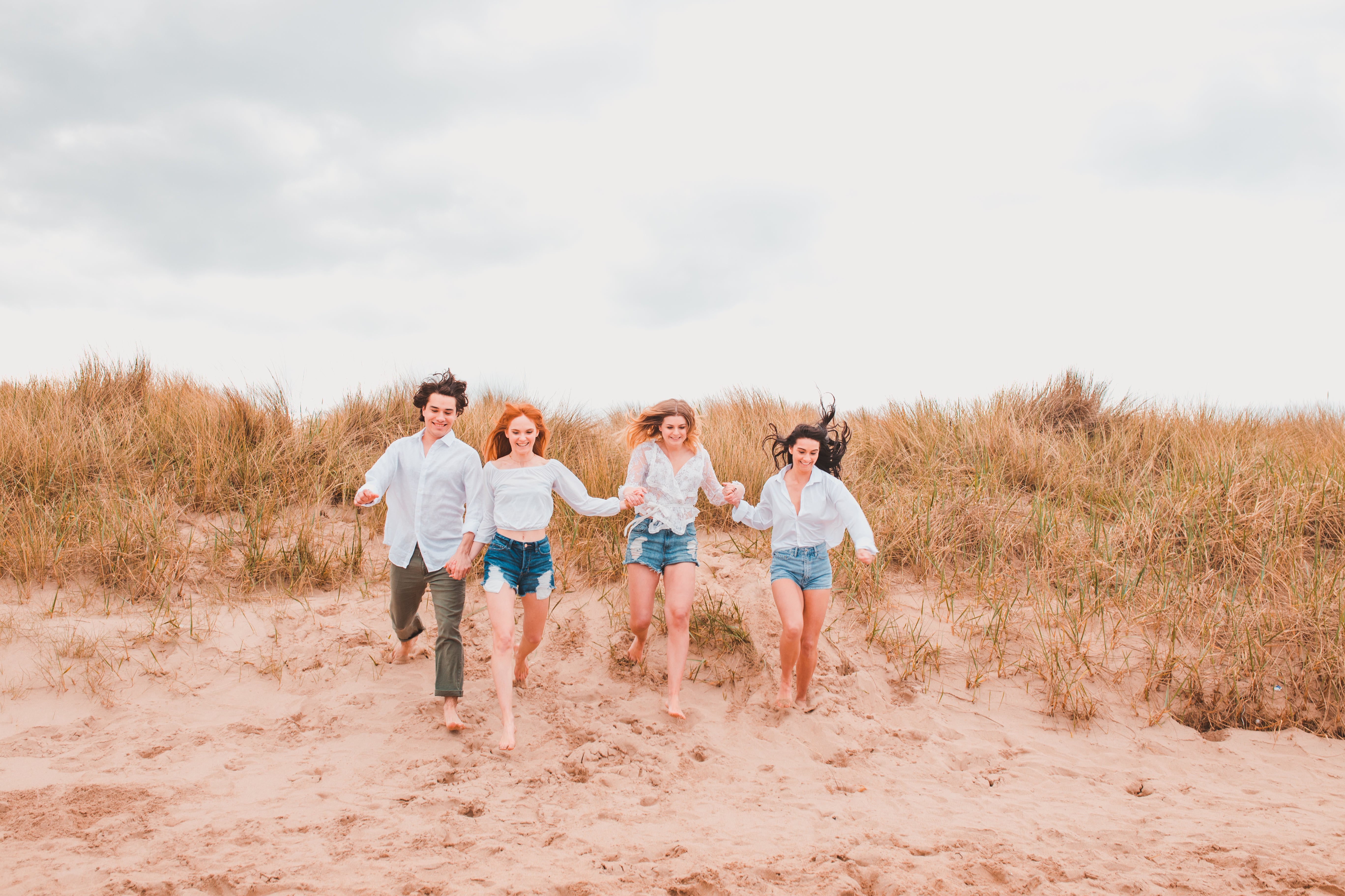 4 people (1 male and 3 females) are all holding hands and running down a sand dune. They are all smiling and laughing. There is a cloudy sky and some dune grass behind them and they are running down the sand.